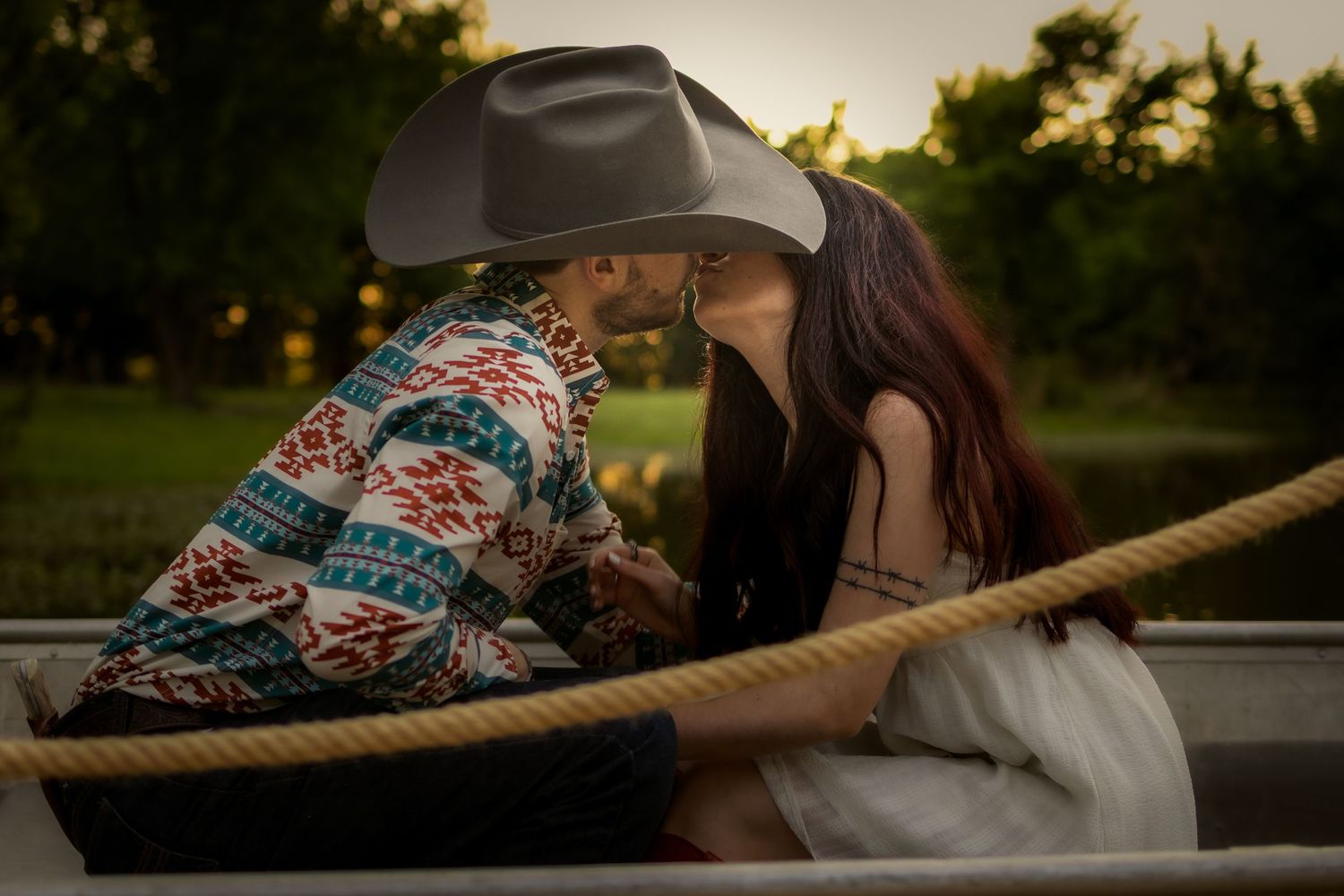 Couple sharing a romantic kiss in a boat at sunset, man wearing cowboy hat and colorful aztec jacket.
