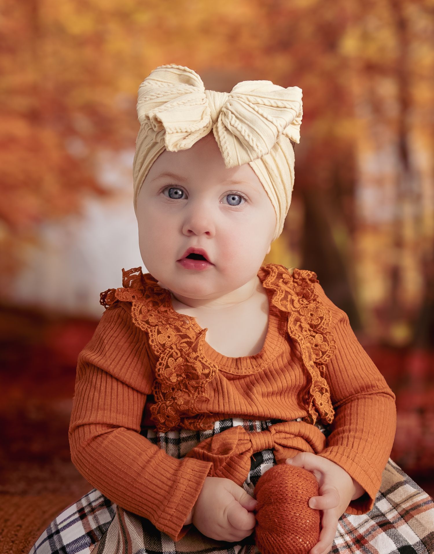 Adorable baby girl in rust lace top and plaid skirt with cream bow headband, posed before fall foliage backdrop.