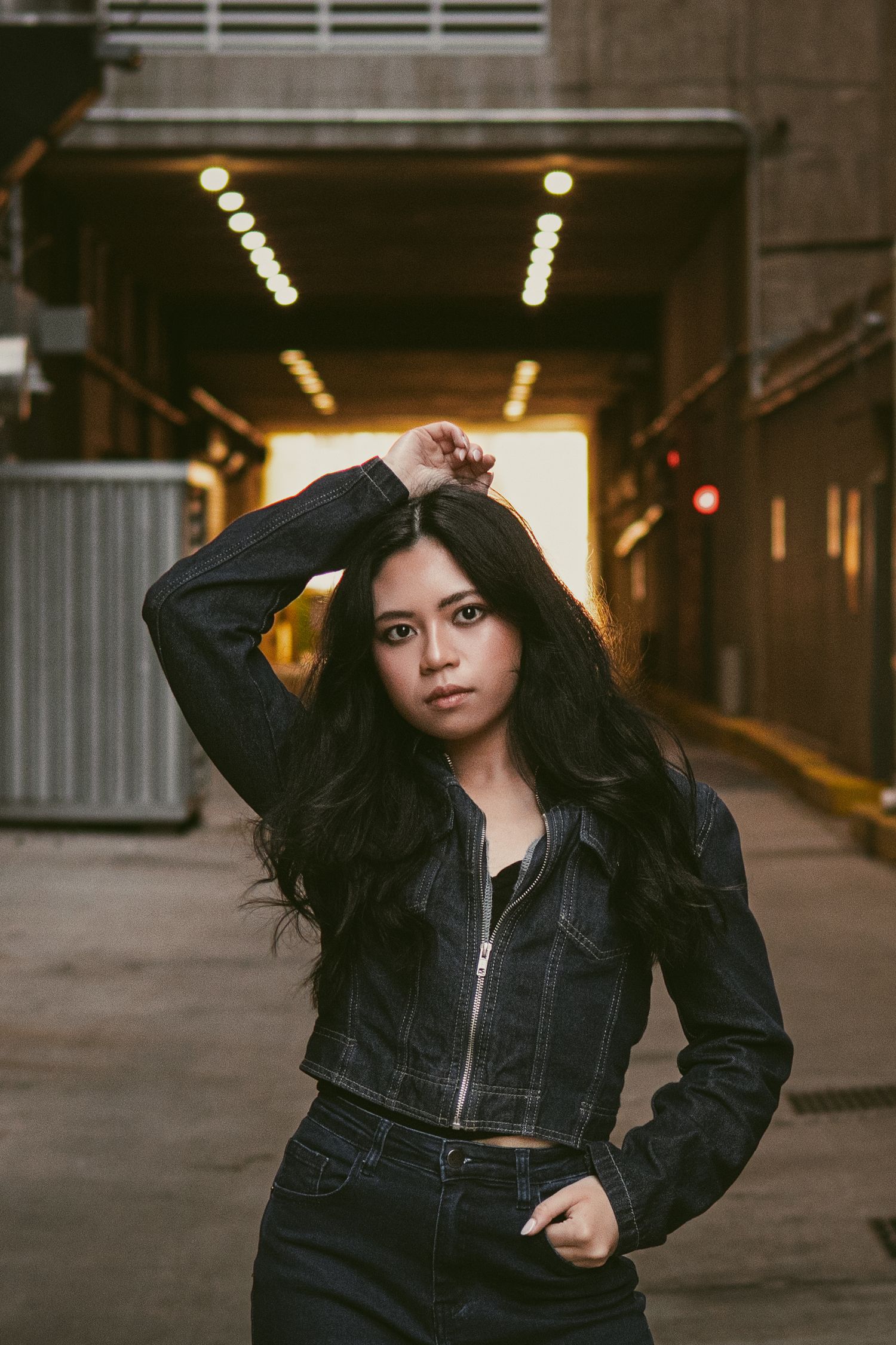 Young woman in denim jacket poses confidently in urban tunnel with bokeh lights background.