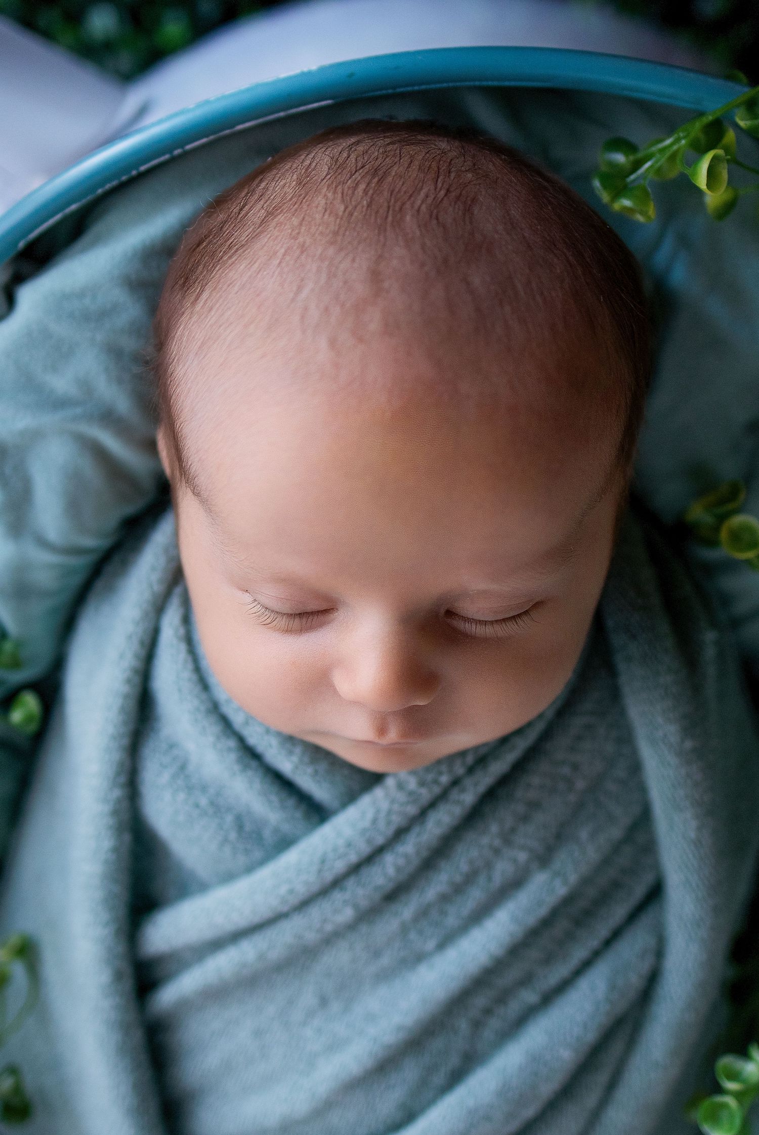 Sleeping newborn baby swaddled in a soft teal wrap, nestled in a matching teal bowl with green leaves.