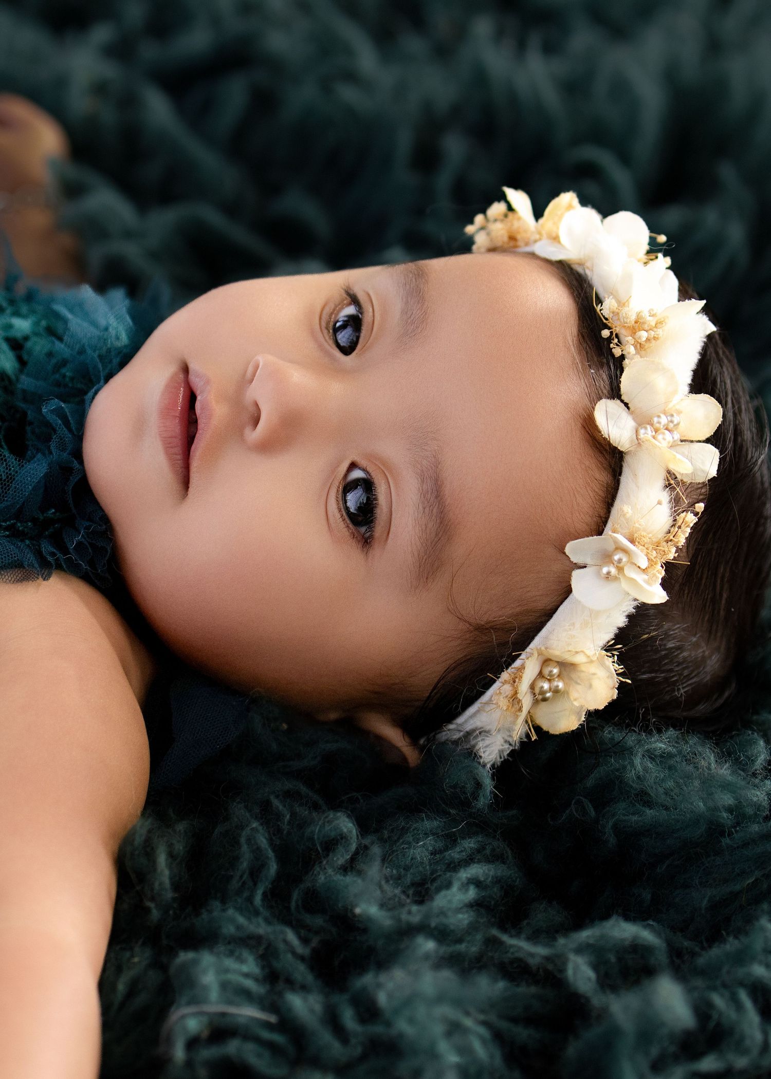 Baby girl wearing a floral crown lying on teal fur, gazing upward with big dark eyes.