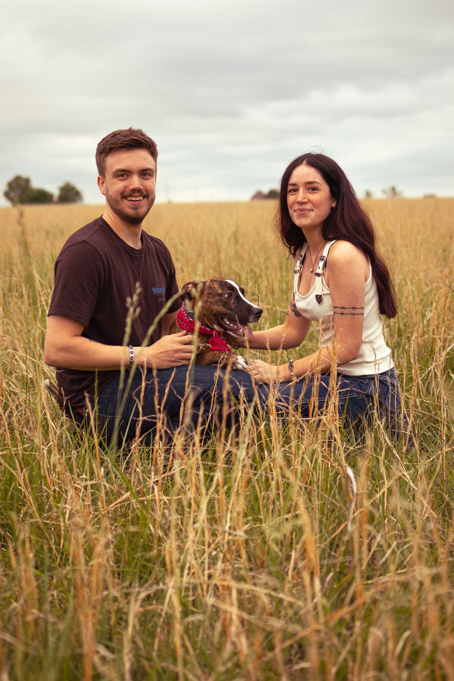 Couple sitting in tall golden grass field with their brown dog wearing a red bandana on a cloudy day.