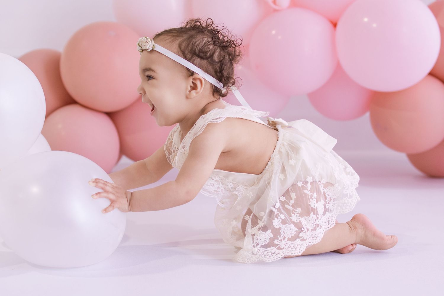 A baby girl in a white lace romper and floral headband plays with balloons at a pink-themed first birthday photo shoot.