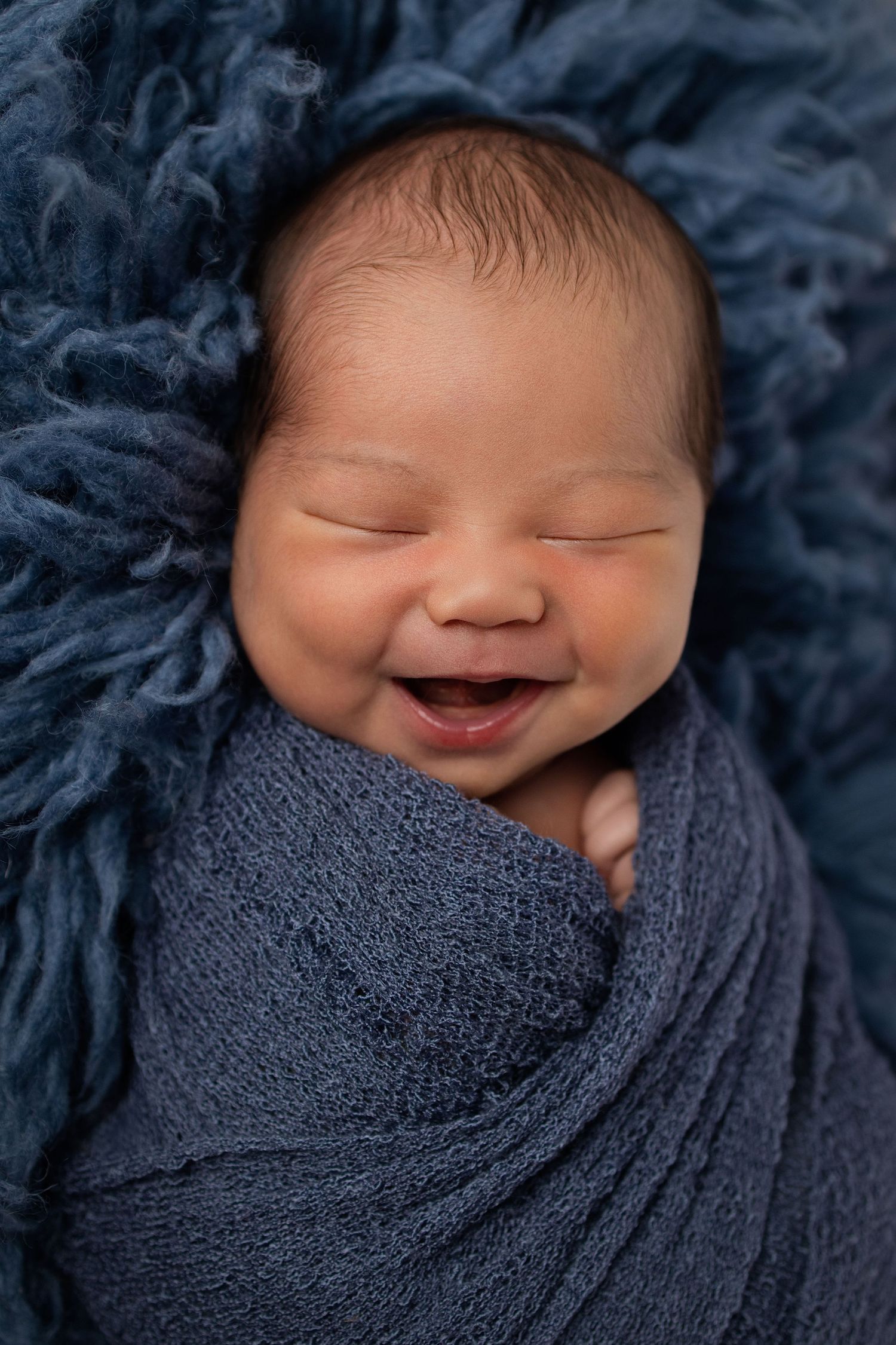 Smiling newborn baby swaddled in a blue wrap, lying on a soft blue fluffy blanket.