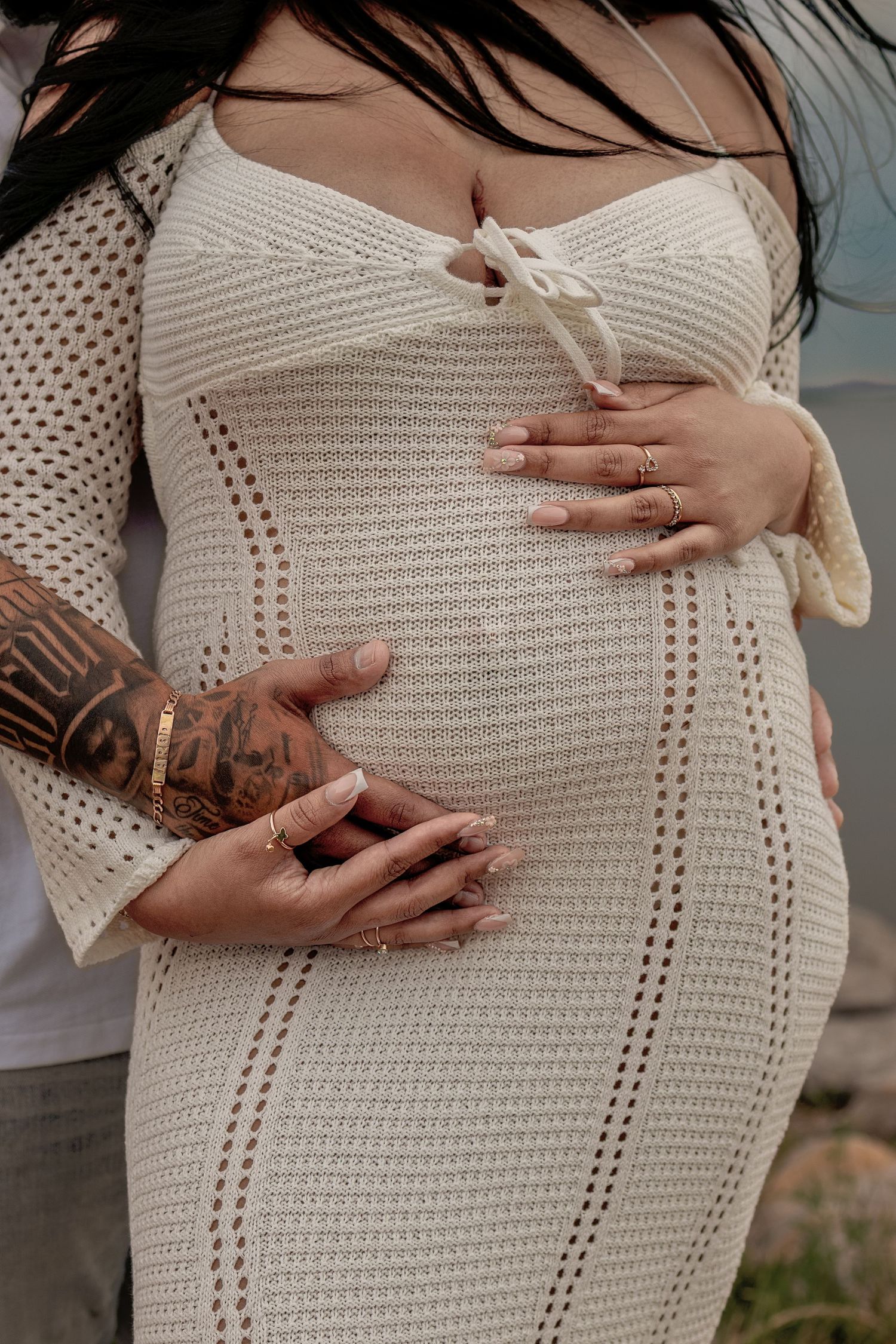 Pregnant woman in cream knit dress with tattooed partner's hands cradling baby bump, maternity photo.