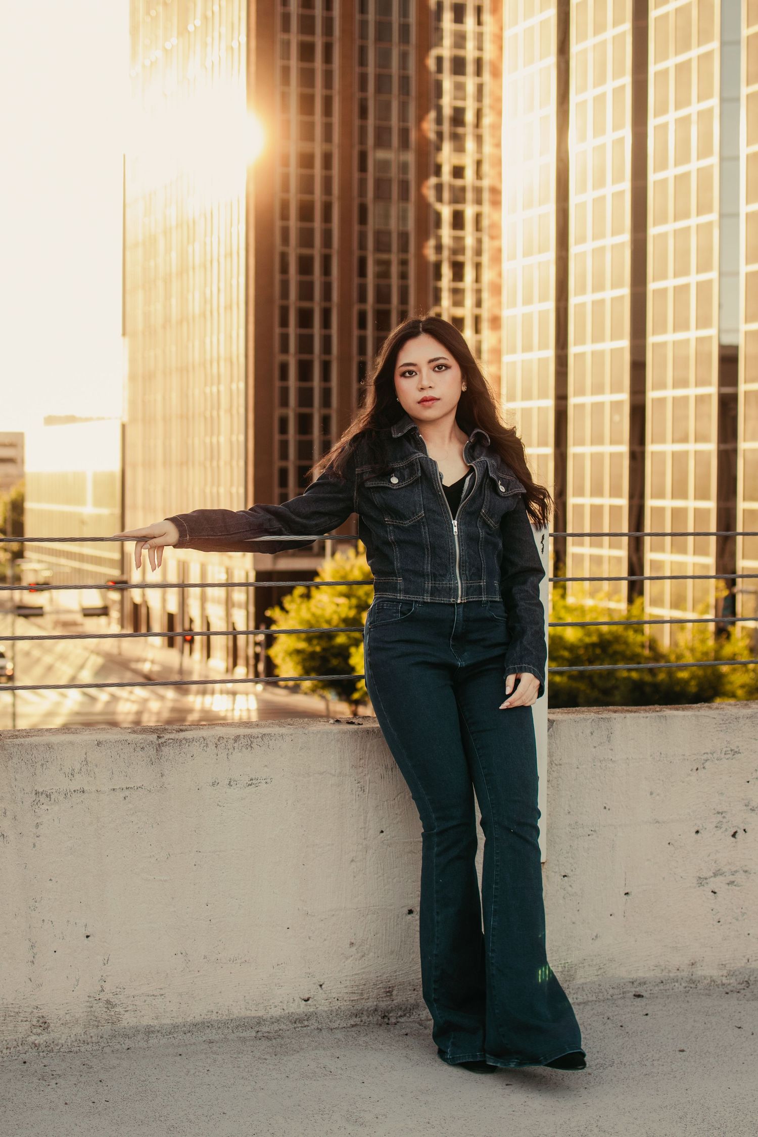Young woman in black leather jacket and flare jeans poses against concrete wall with golden hour city skyline backdrop.