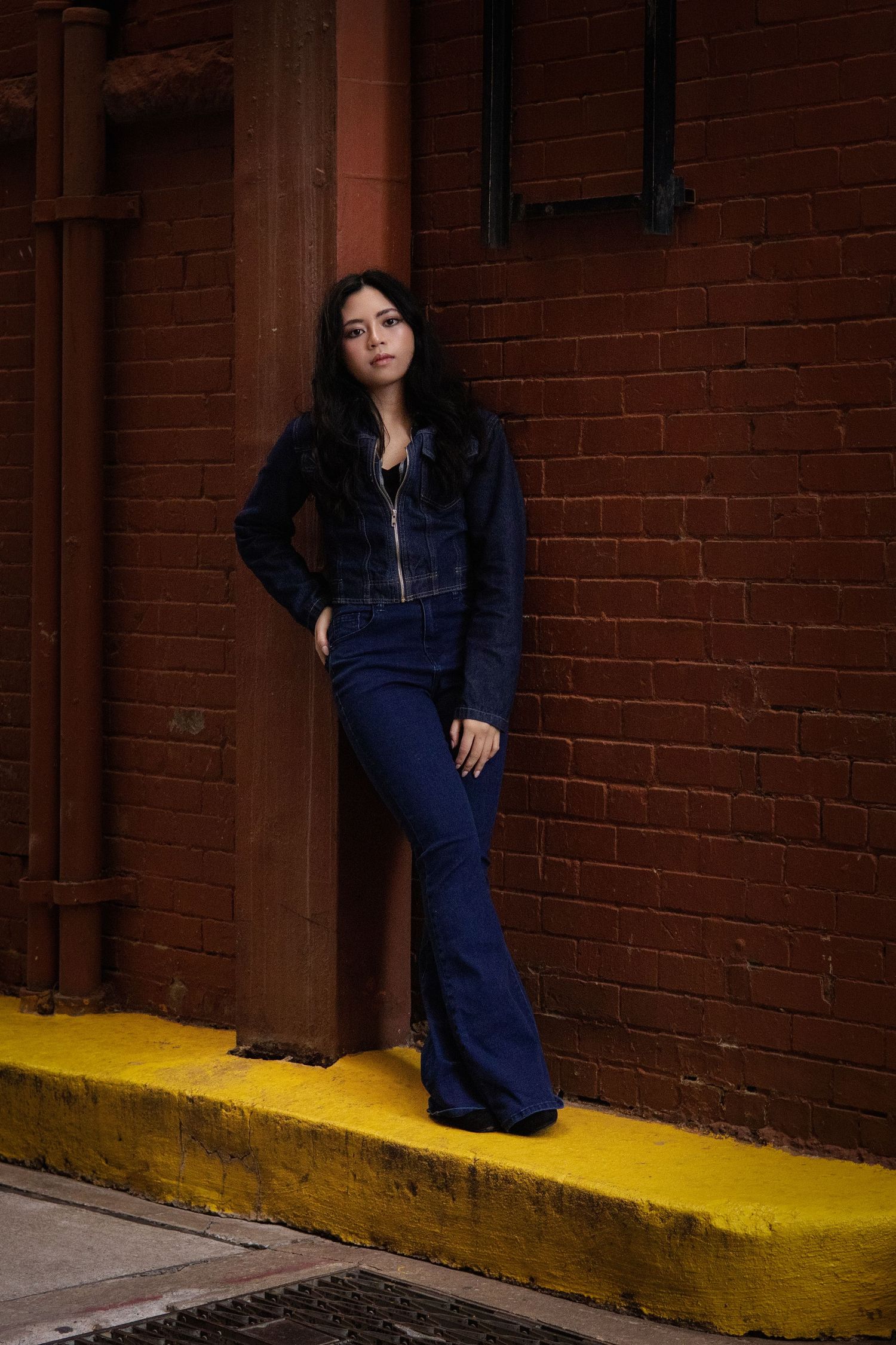 Young woman in denim jacket and flare jeans leans against brick wall on yellow curb in moody urban alley.