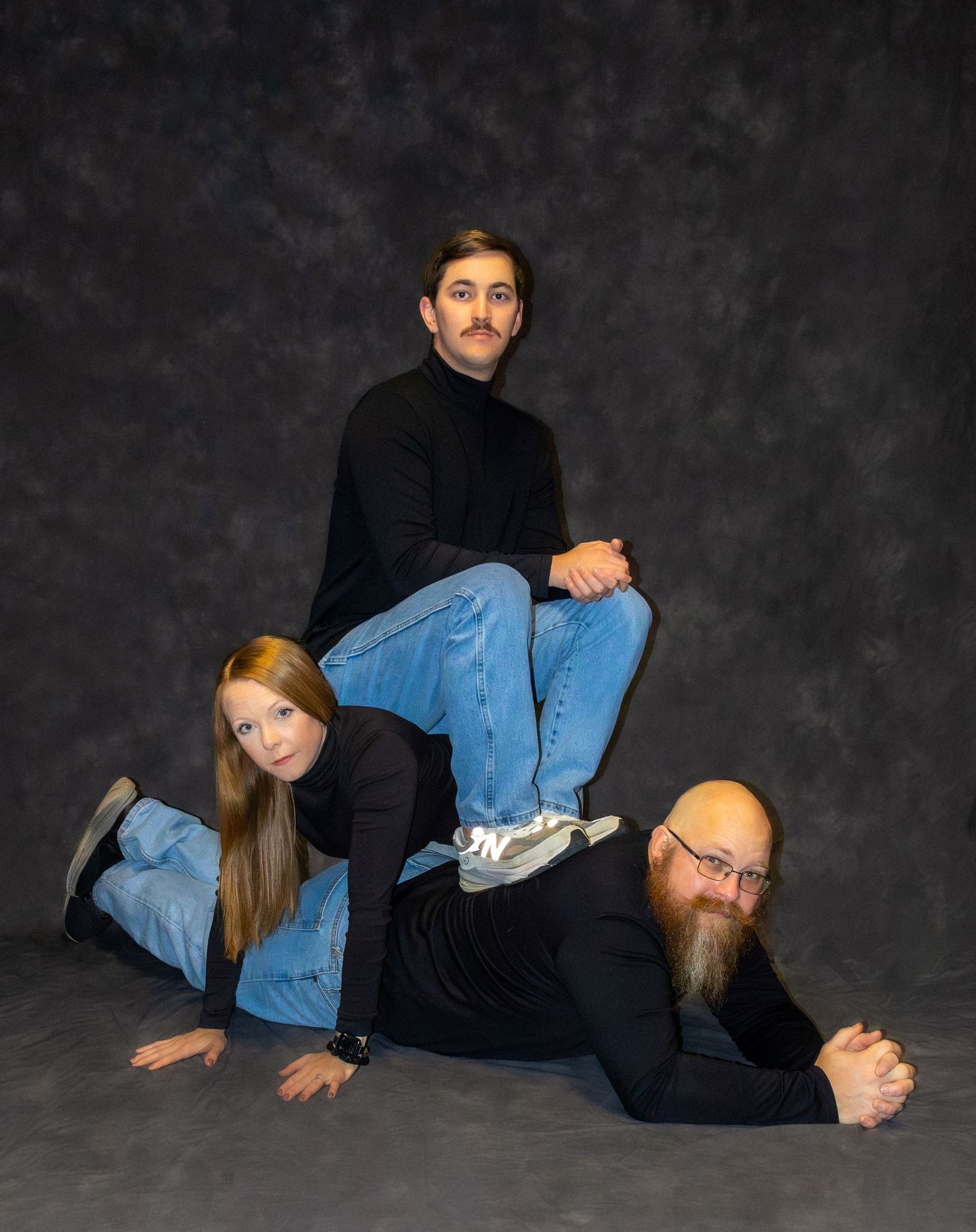 Three people in black outfits pose creatively in a studio, with one sitting atop the others in a playful pyramid formation.