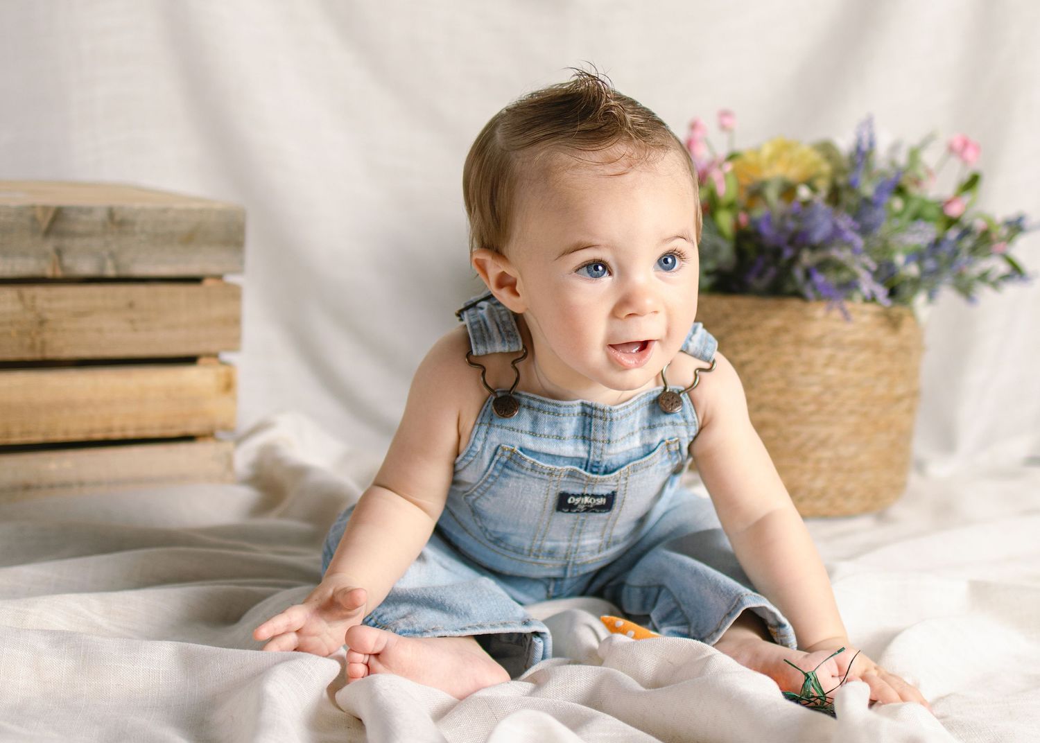 Cute baby in denim overalls sitting on white blanket smiling with flower basket behind them.