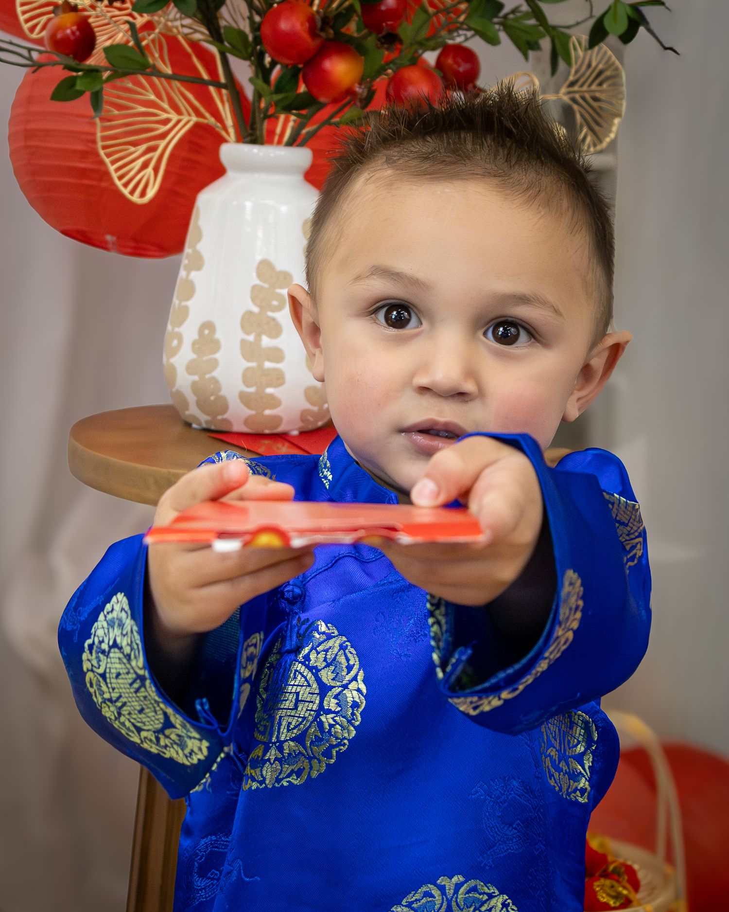 Young boy in blue gold brocade ao dai holds red envelope, with lanterns and flowers in background.
