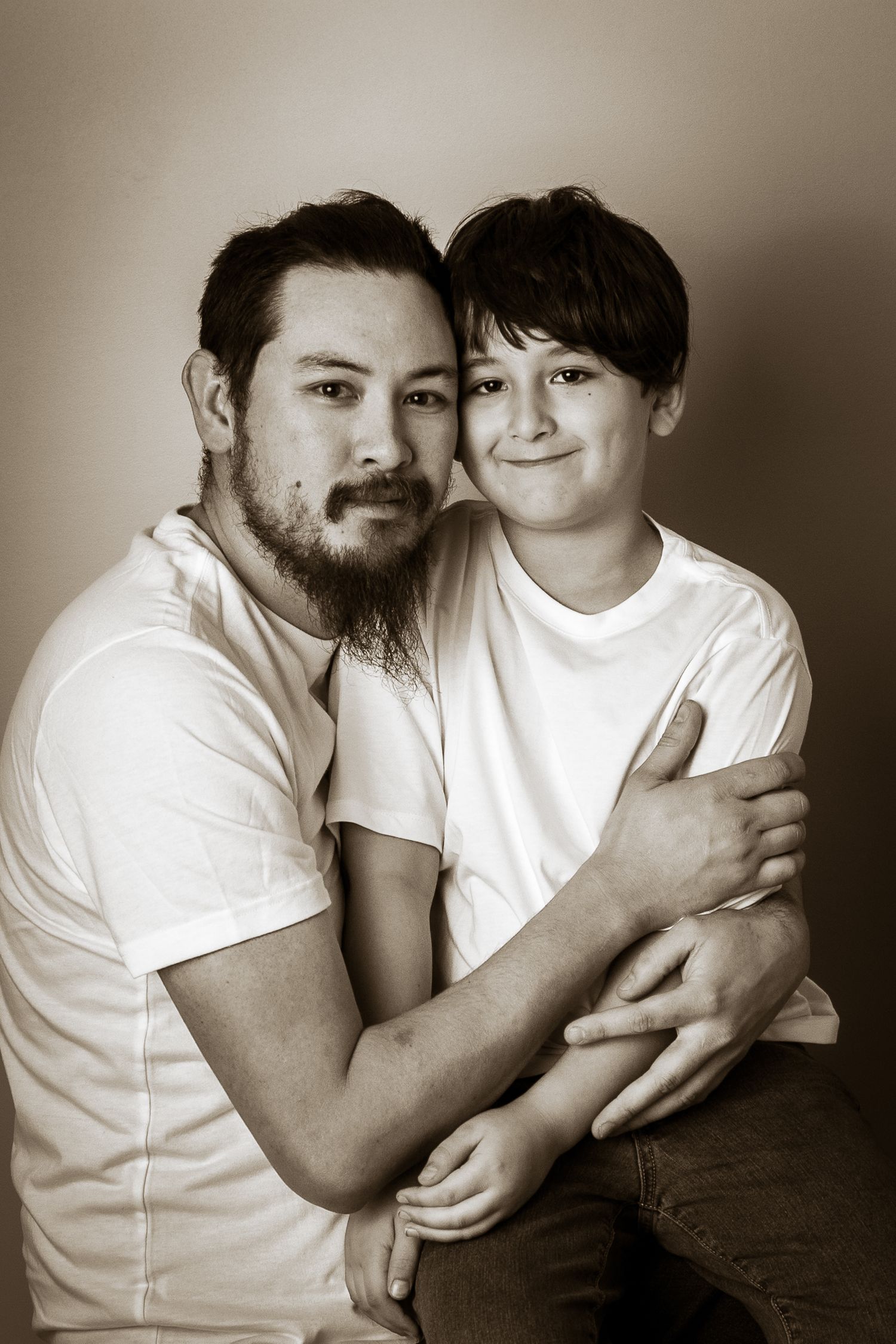 Sepia-toned portrait of a bearded man and young boy in white shirts sharing a warm embrace.
