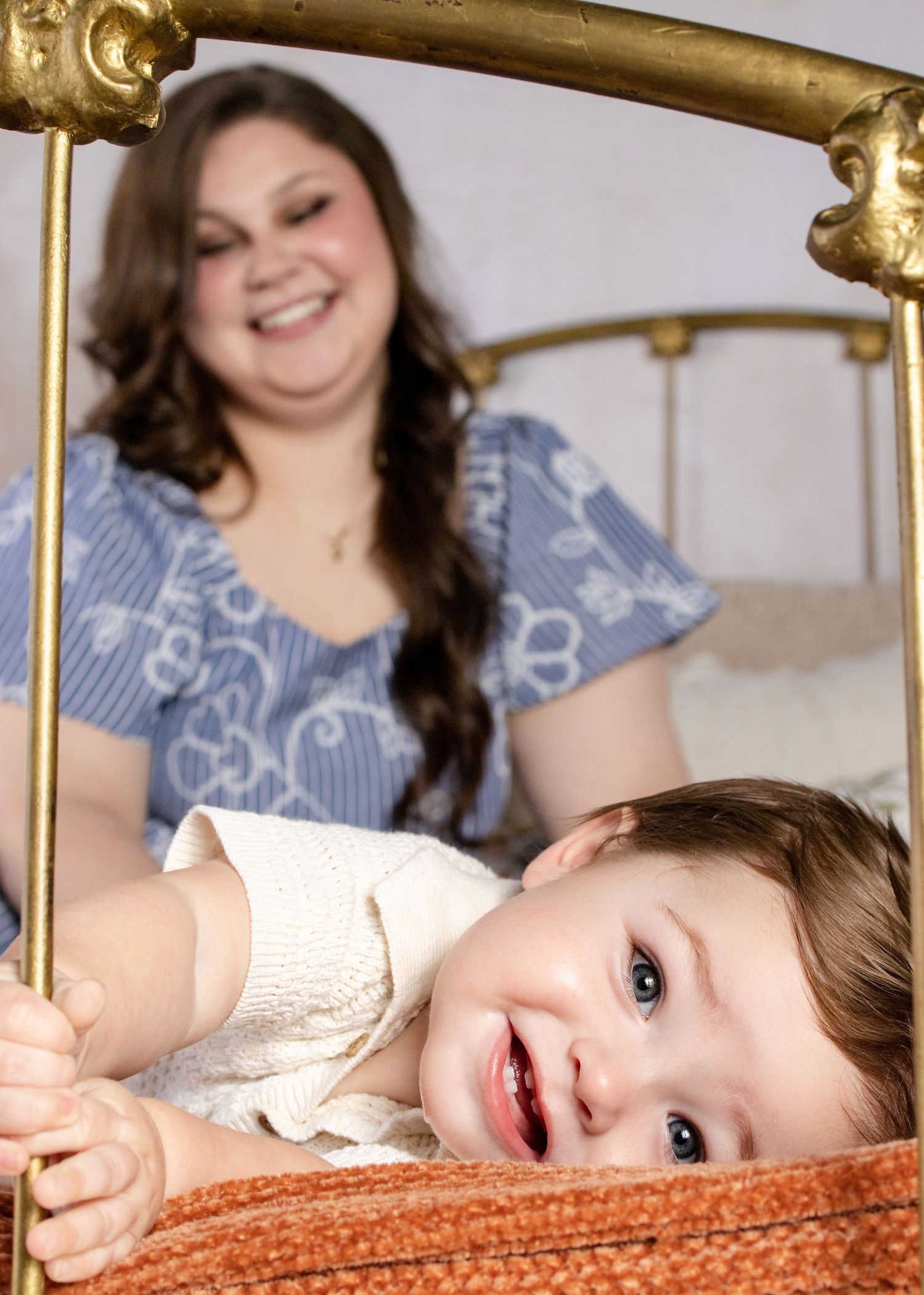 A smiling baby leans on a gold bed frame while a happy young woman smiles in the background.
