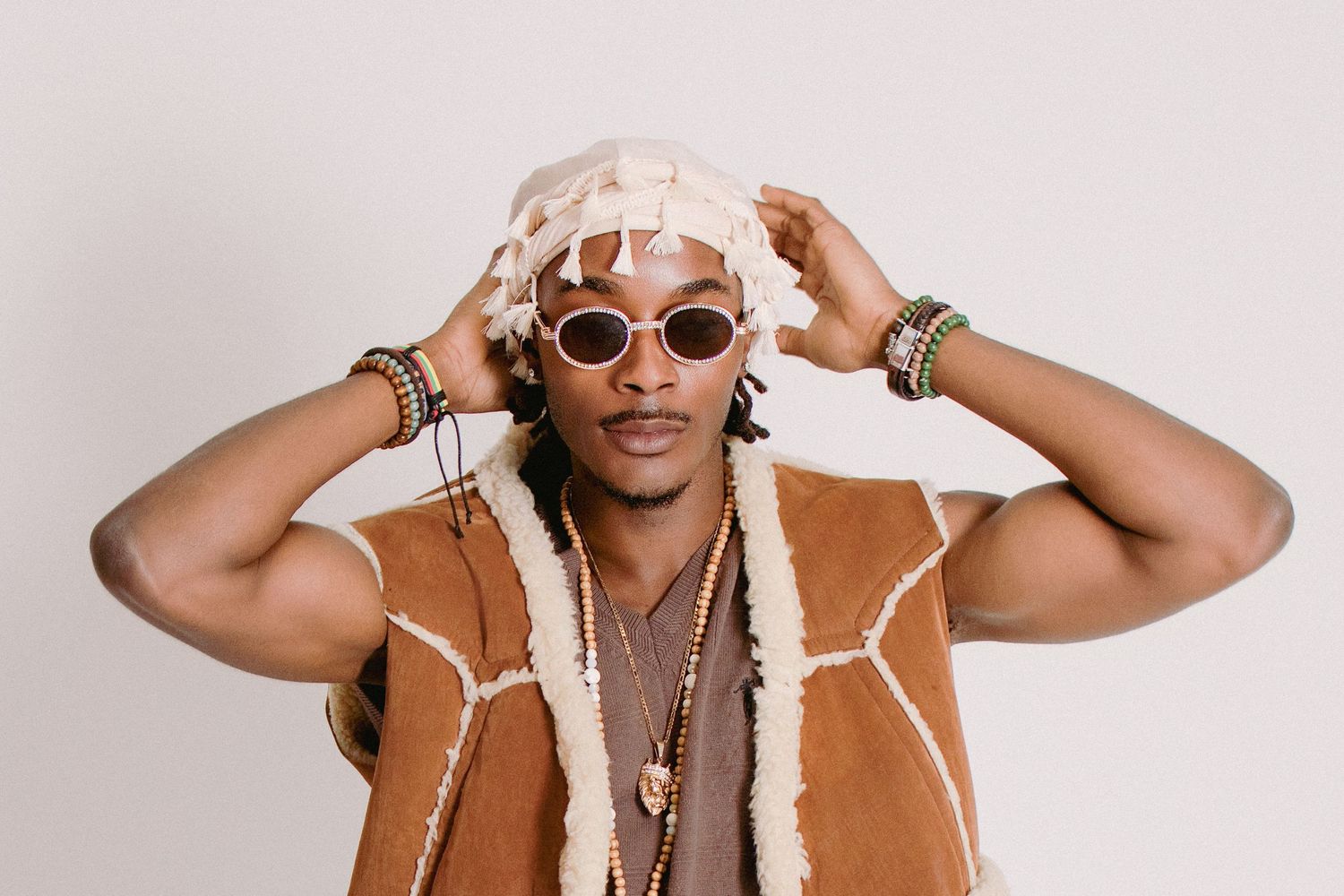 Stylish man wearing pink sunglasses, beaded necklaces, suede vest, and a fringed hat against a light background.