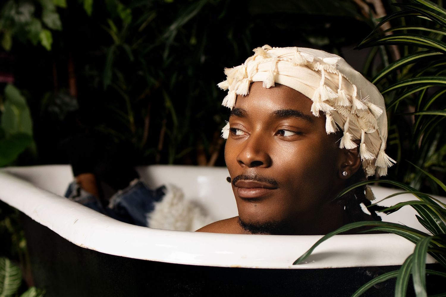 Young Black man wearing white shower cap relaxes in clawfoot bathtub surrounded by lush green plants.
