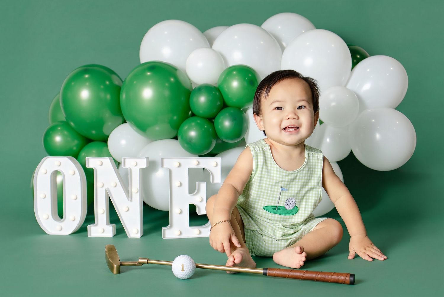 Baby's first birthday photo with green balloons, ONE marquee letters, and golf club props on green background.