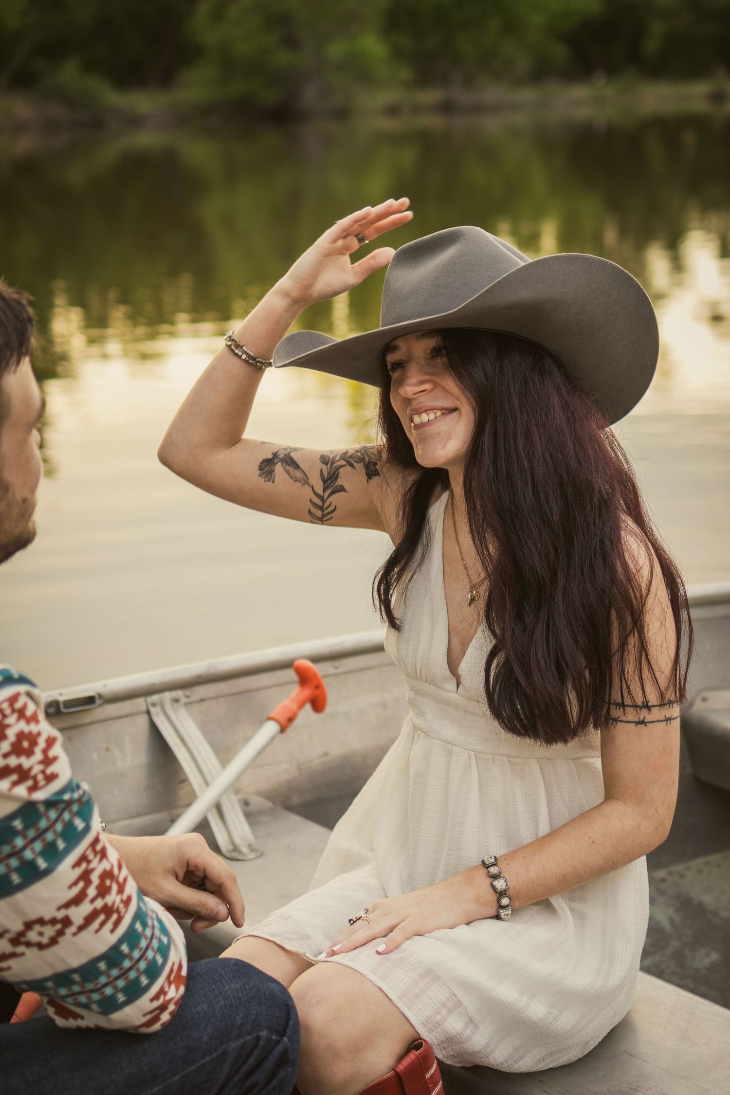 Smiling woman in white dress and wide-brim hat sitting in a canoe on a lake, showing arm tattoos.
