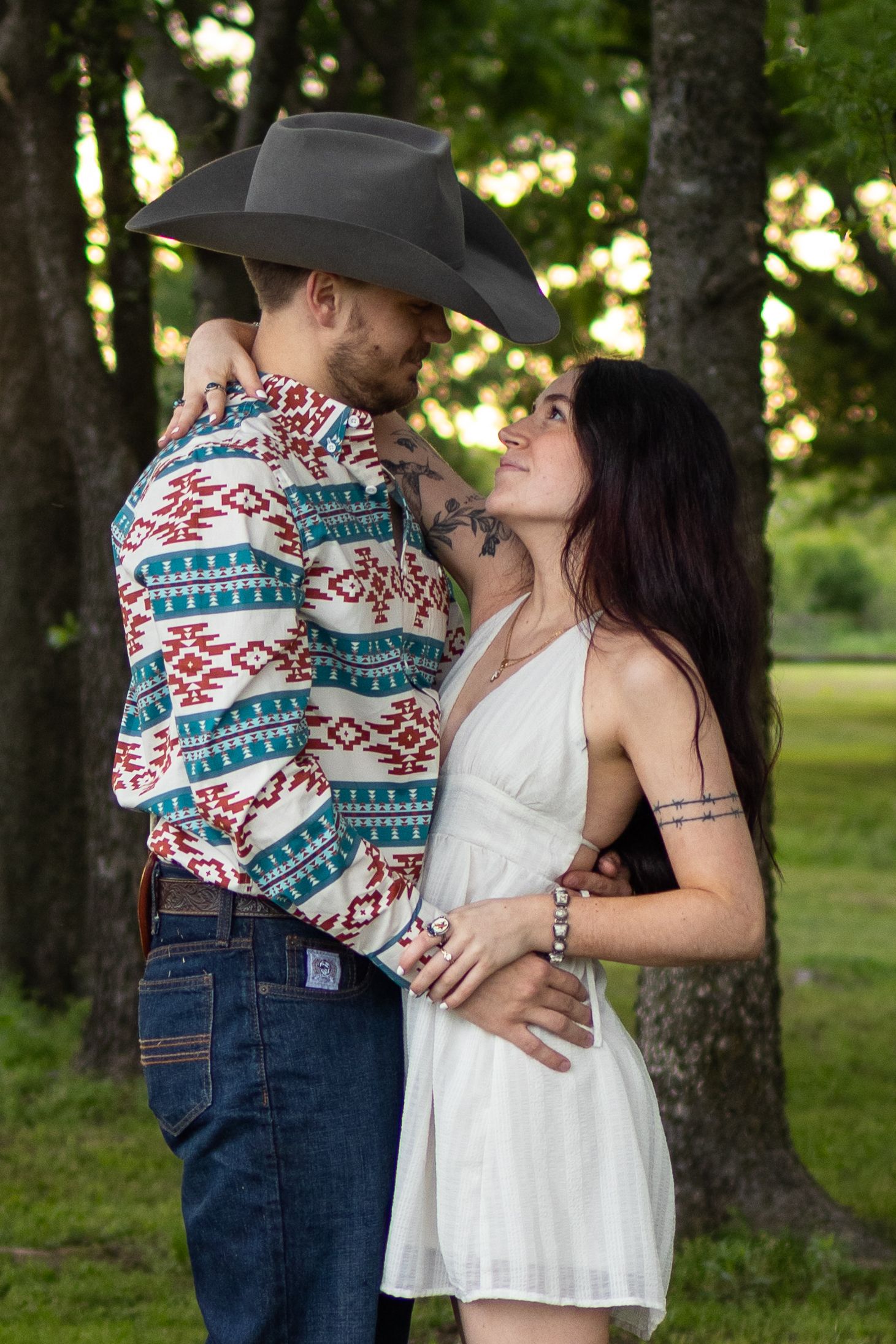 Couple embracing outdoors, man in cowboy hat and patterned shirt, woman in white dress.