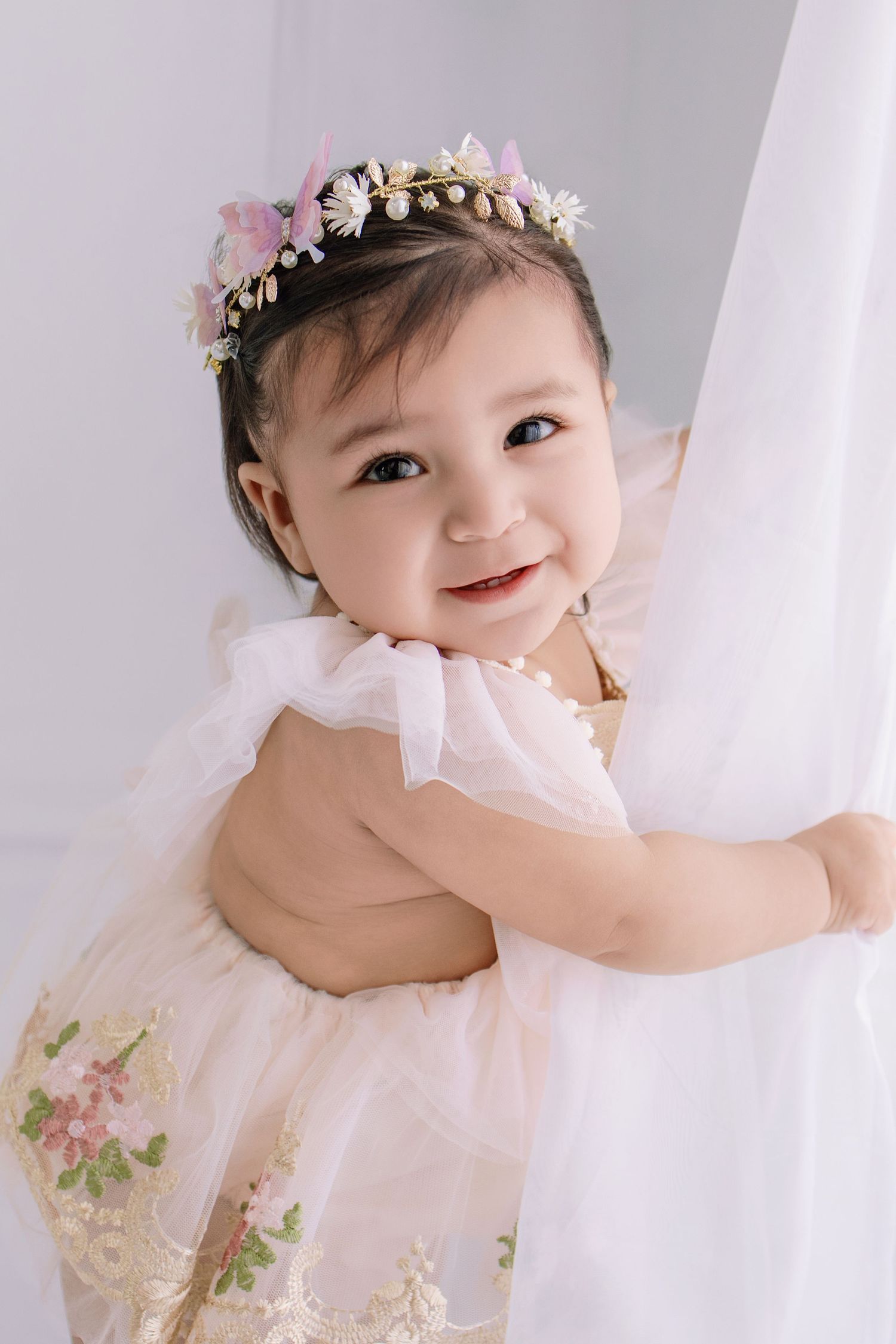 Smiling baby girl in a floral crown and pink embroidered tulle dress, holding a white curtain in a bright studio.