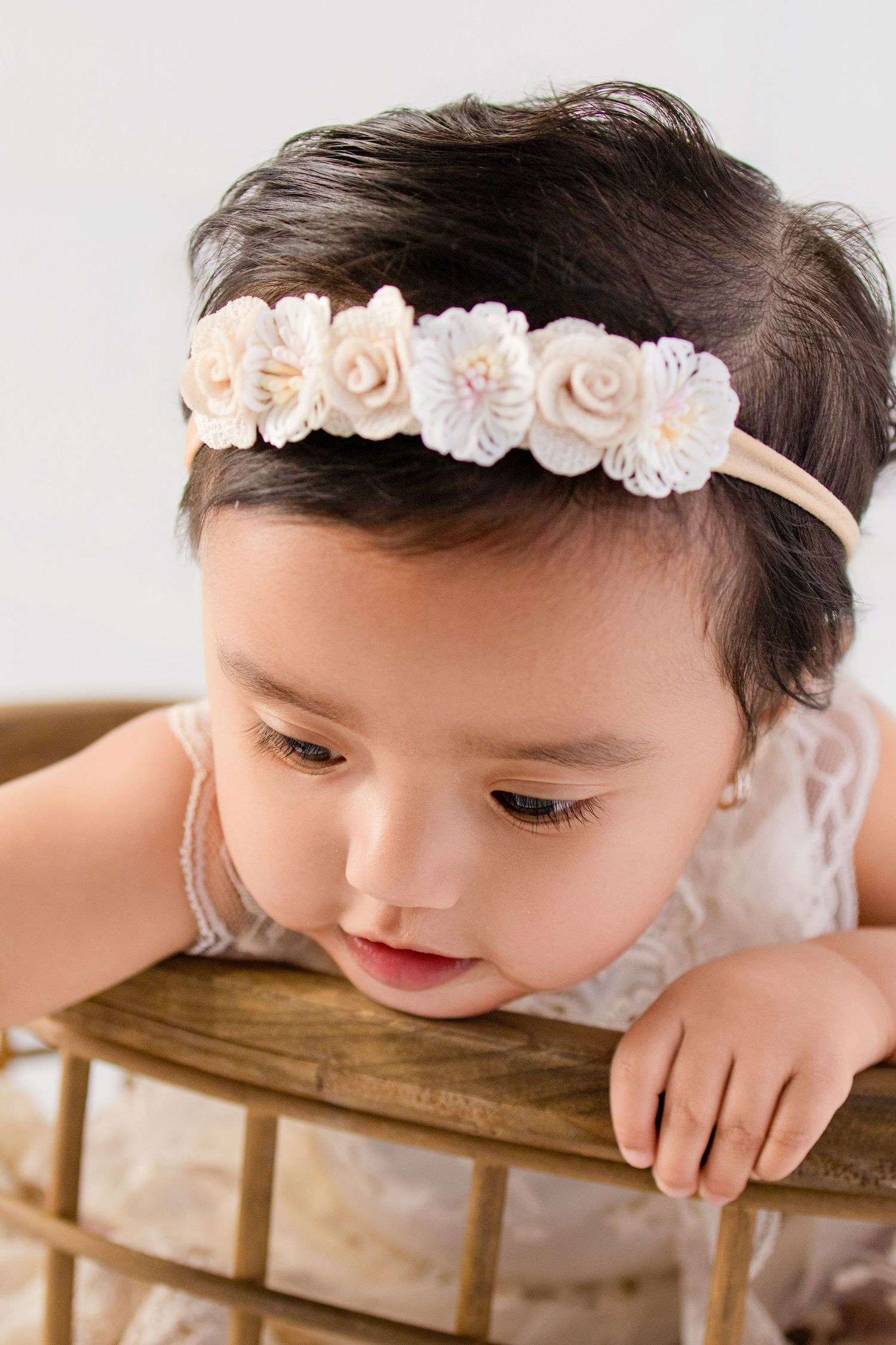Baby girl wearing a delicate floral headband, leaning on a wooden chair in a soft white studio setting.