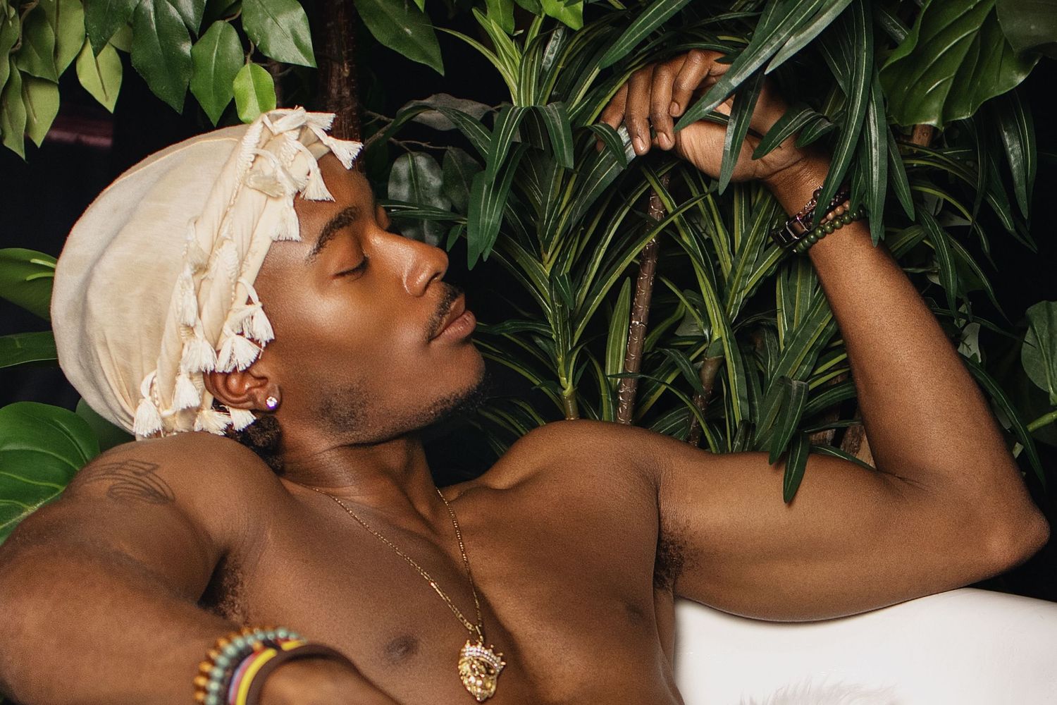 A shirtless man with a head wrap relaxes among lush tropical green plants wearing beaded jewelry.