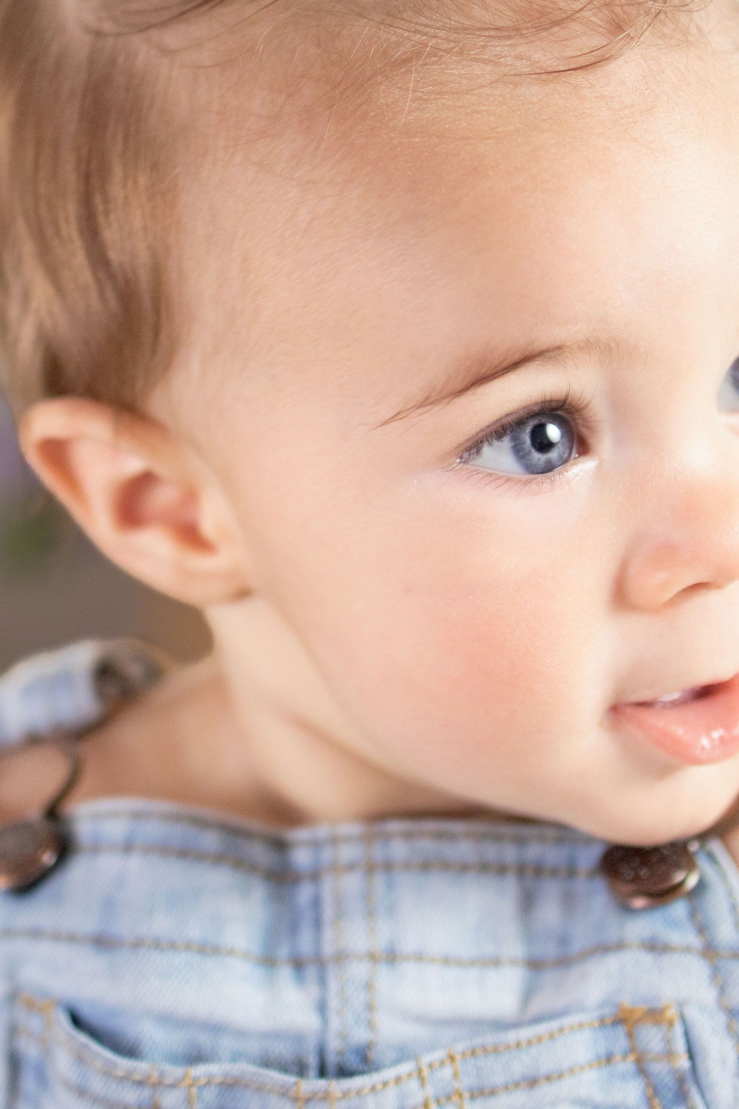 Close-up portrait of a blue-eyed baby in denim overalls looking to the side.
