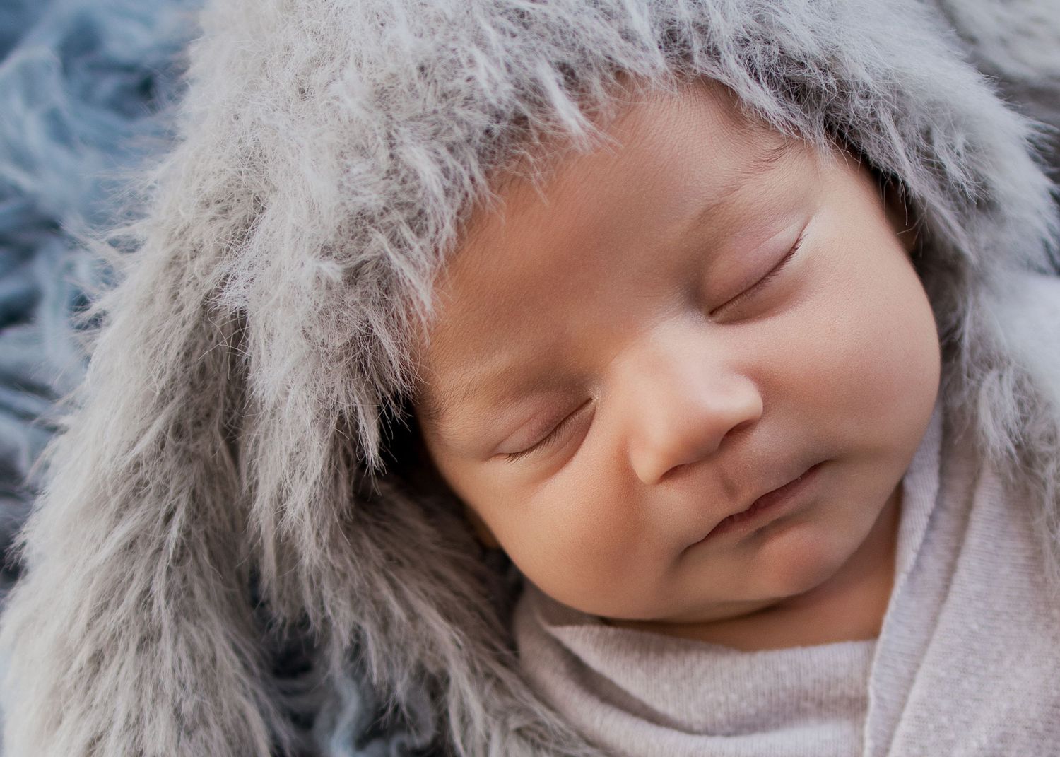 Adorable newborn baby sleeping in a fluffy gray hat on a soft blue blanket.
