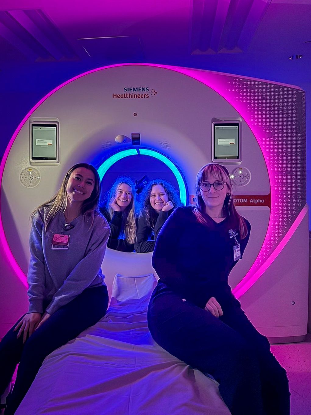 Four women posing in front of a Siemens Healthineers MRI machine with colorful pink and blue lighting.