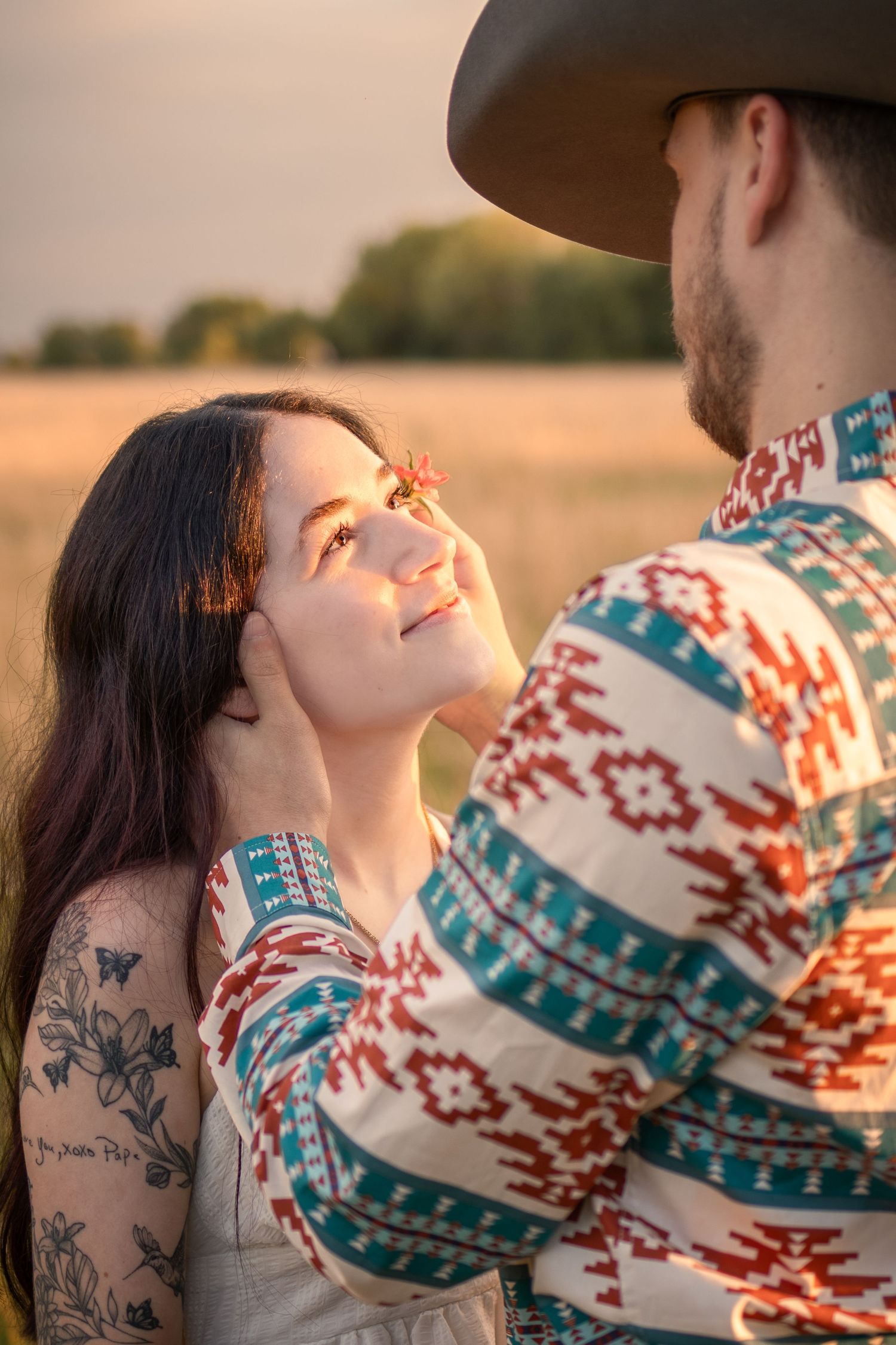 A couple shares a romantic moment at golden hour in a field, the man in a southwestern shirt and cowboy hat gazing at a tattooed woman.