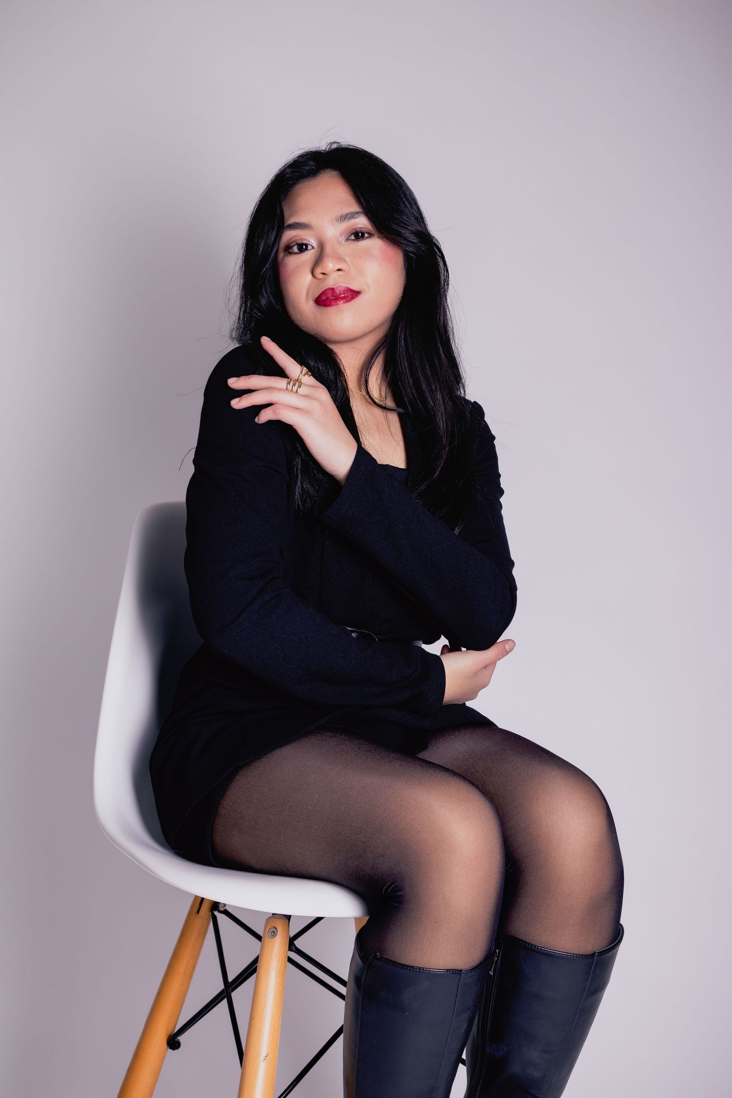 Woman in black dress and boots sitting on white chair posing for studio portrait photo.