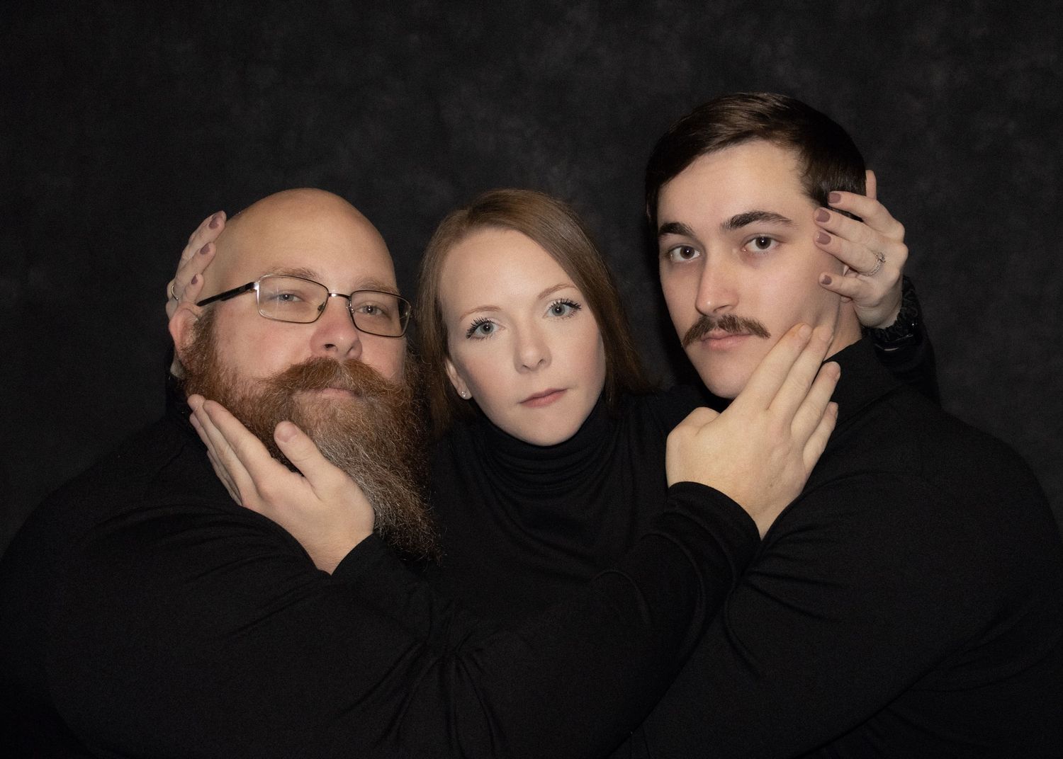 Three adults in black turtlenecks pose dramatically against a dark background, two men flanking a woman.