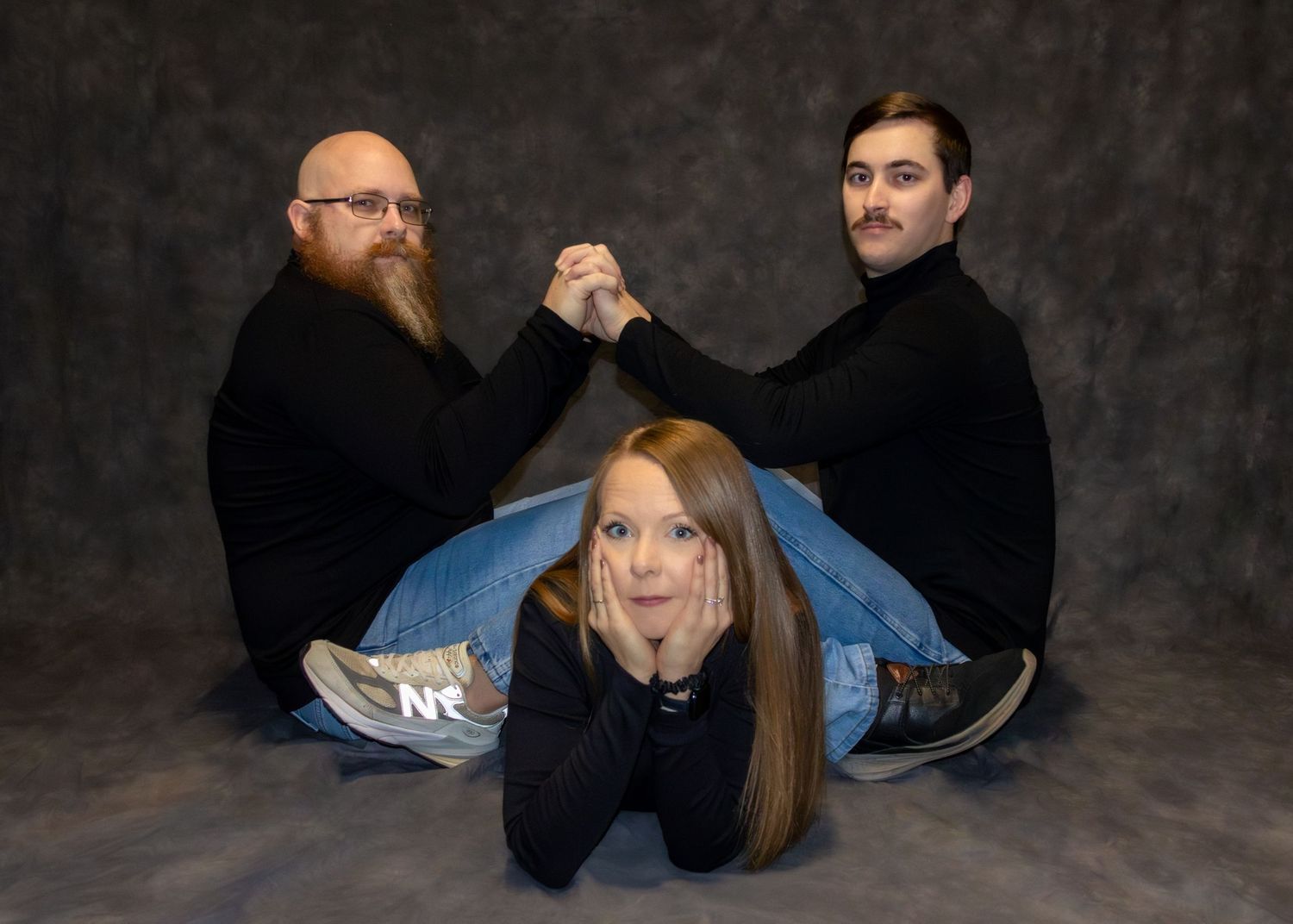 Two bearded men sitting back-to-back arm wrestling while a young blonde woman poses in front on a gray backdrop.