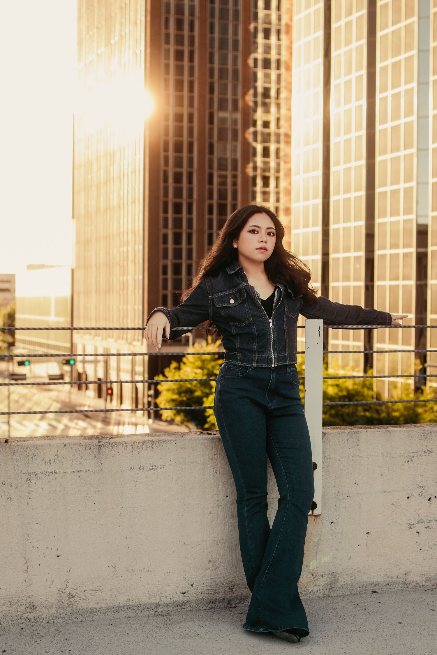 Brunette woman in black denim outfit leans against rooftop railing with sunlit skyscrapers glowing behind her.