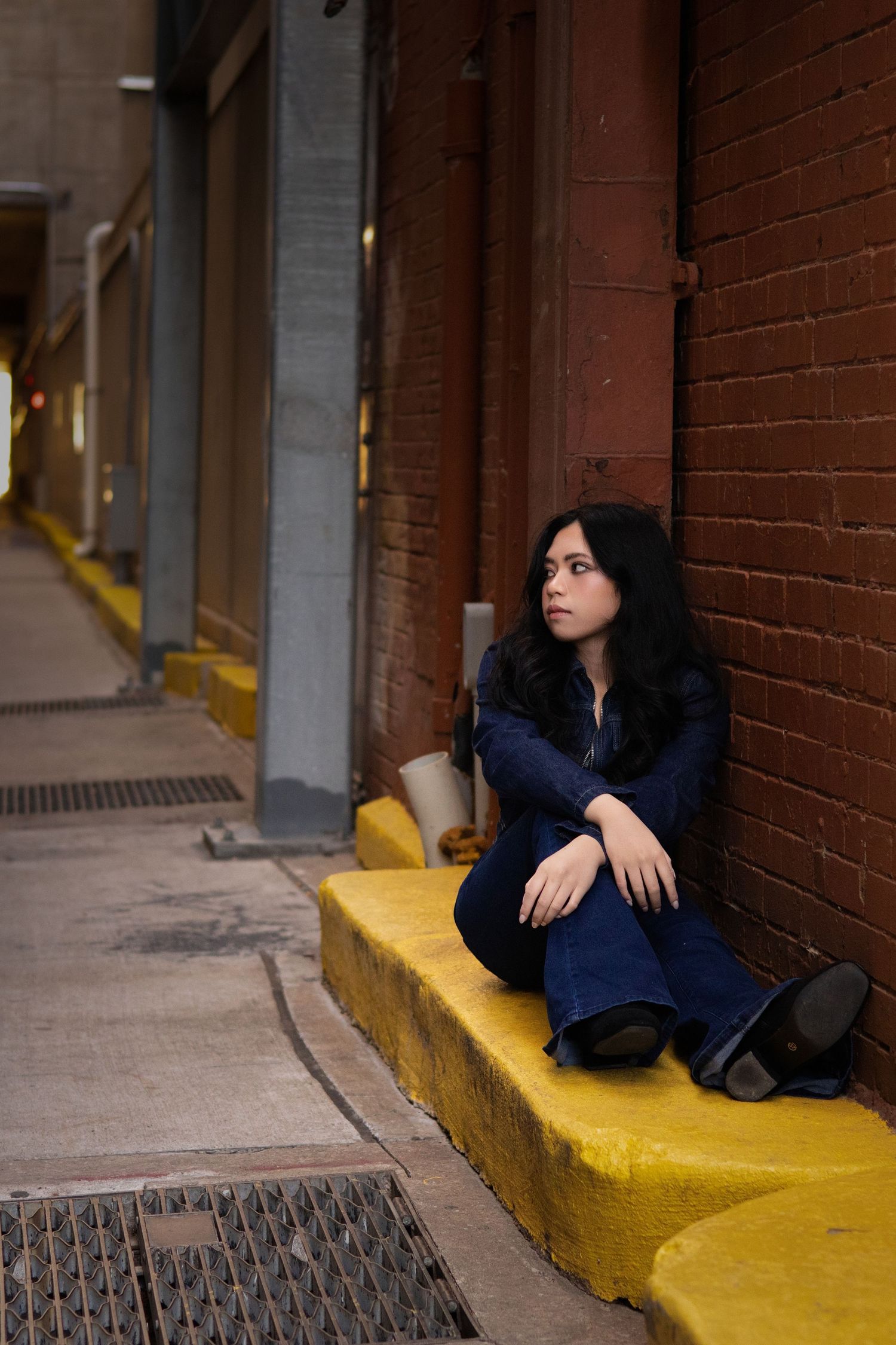 Contemplative young woman in blue jeans and jacket sits on yellow painted curb beside red brick wall in city alley.