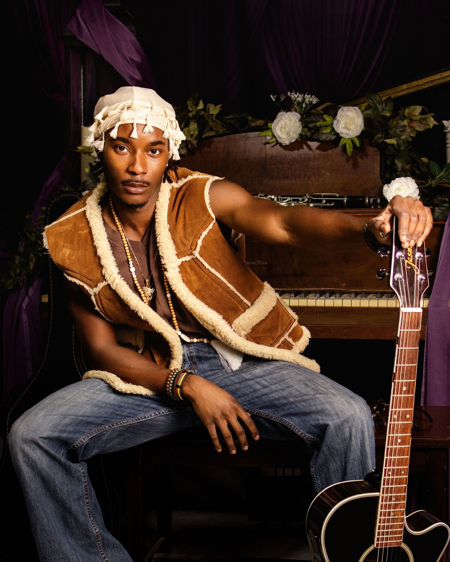 Young man in shearling vest and head wrap poses with acoustic guitar near vintage piano with purple drapes.