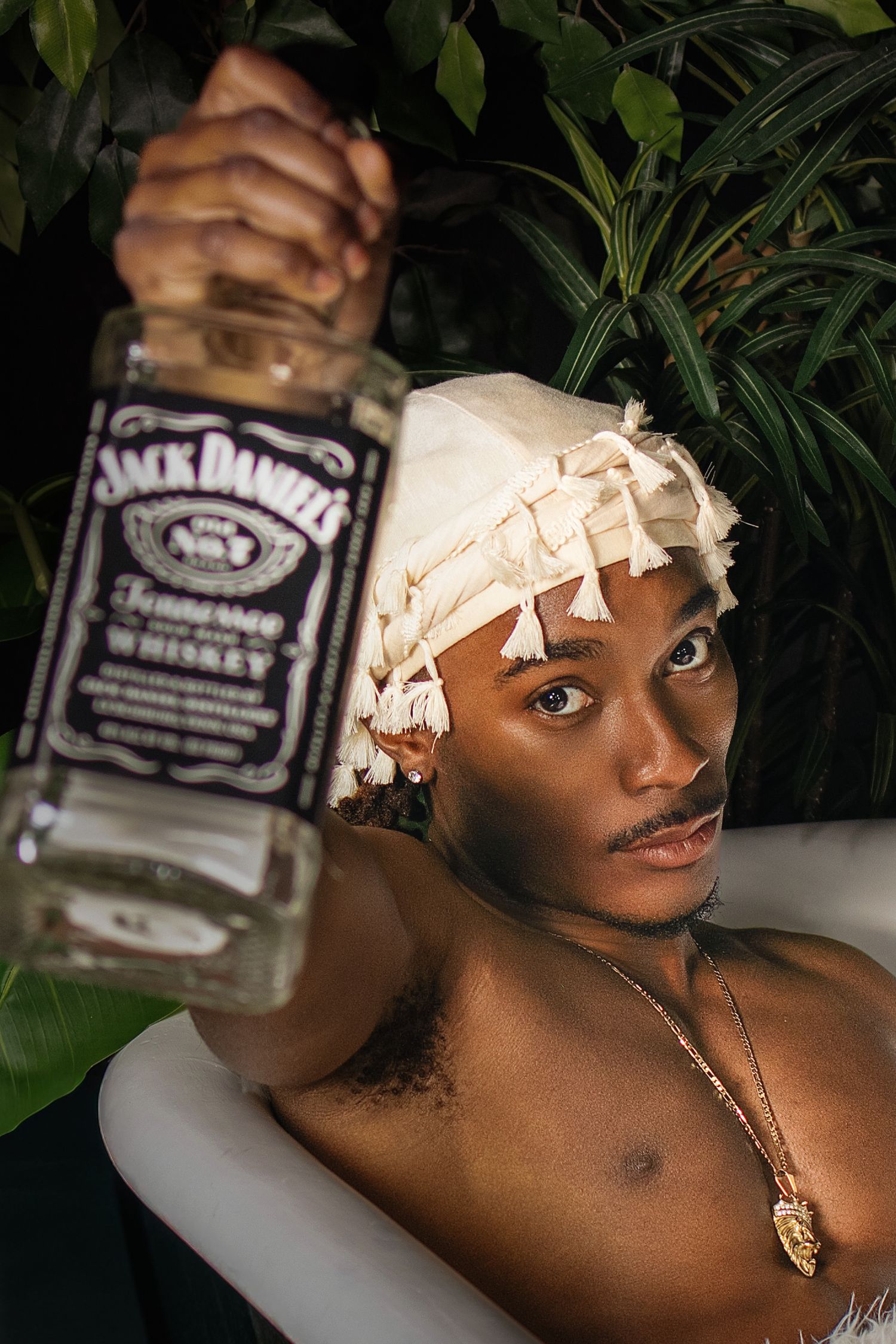 Young Black man in bathtub holds Jack Daniels whiskey bottle wearing decorative head wrap outdoors.