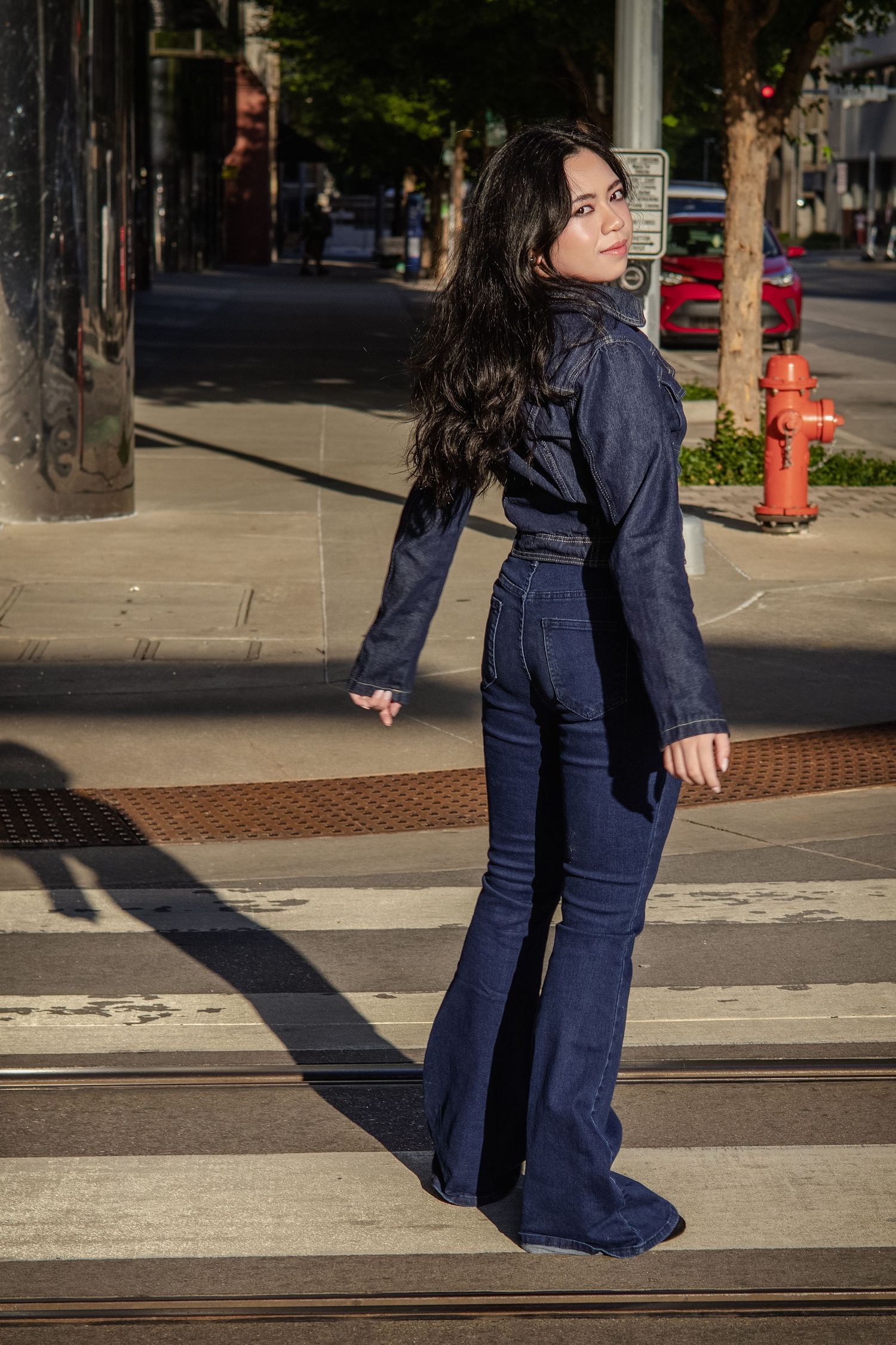 Woman in dark denim flare jumpsuit poses on urban crosswalk with city street and fire hydrant in background.