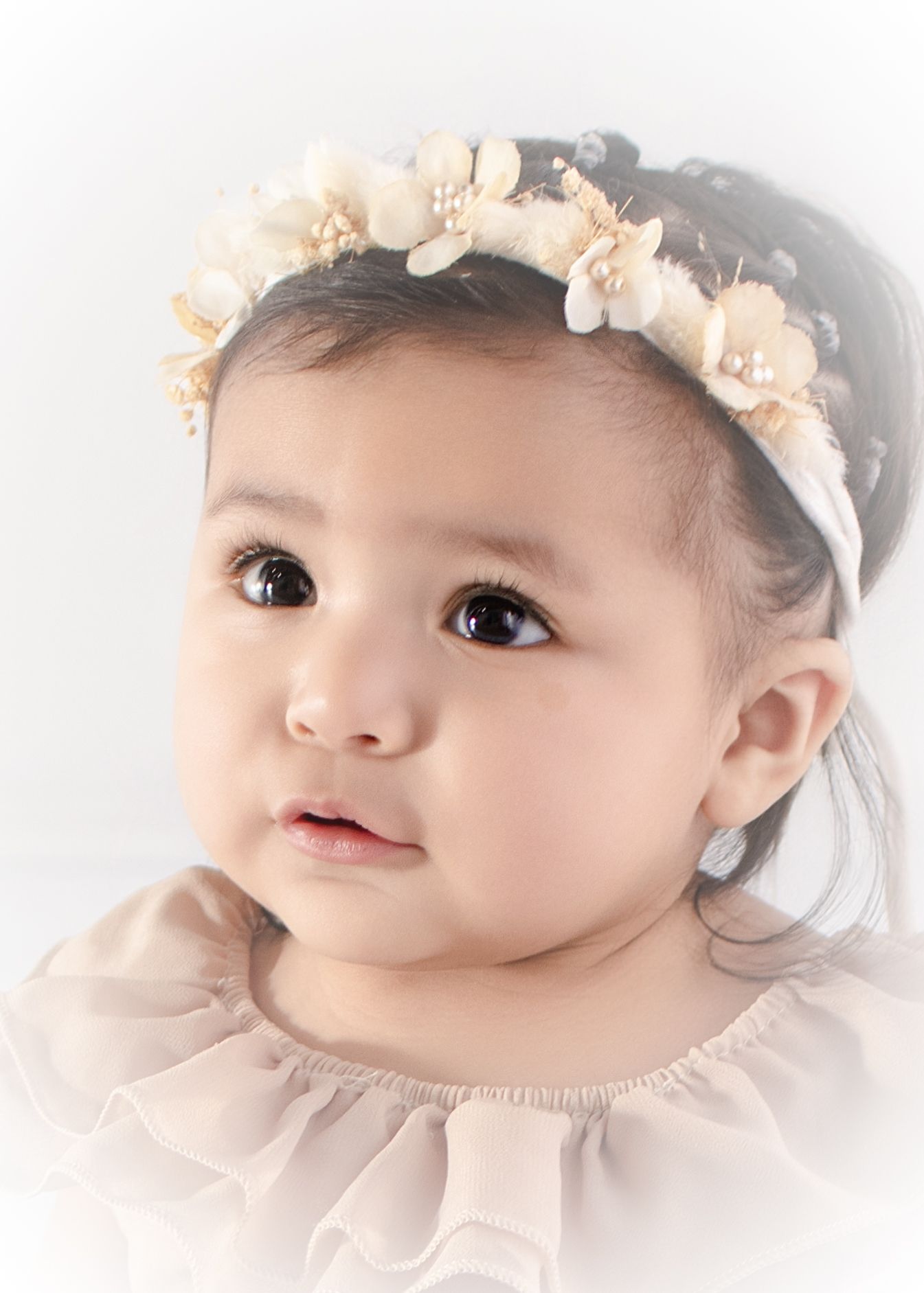 Adorable baby girl wearing a floral headband with pearls, dressed in a soft pink dress, looking up with big brown eyes.