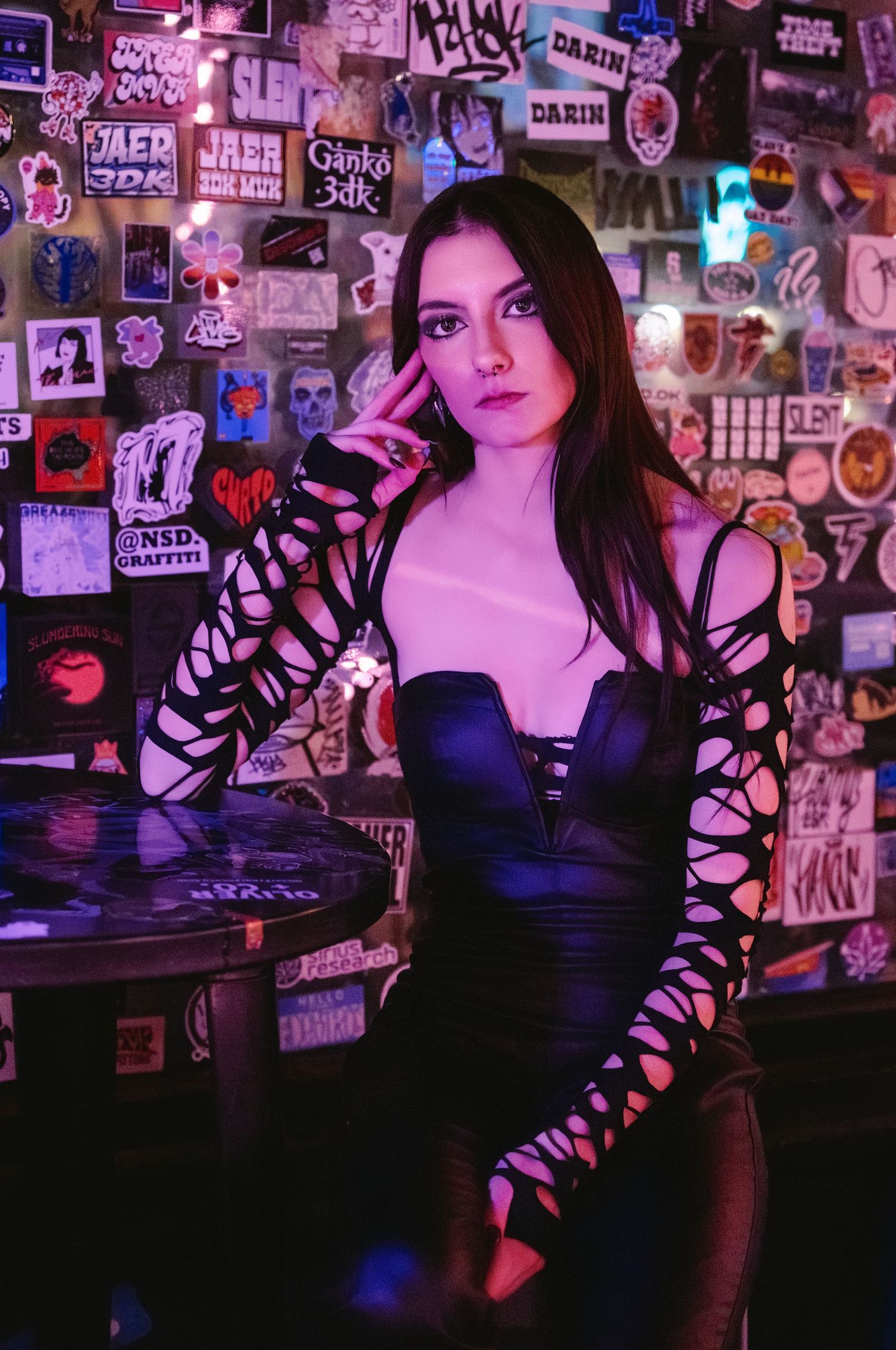 Young woman in black cutout dress poses at table covered in stickers in neon-lit bar.