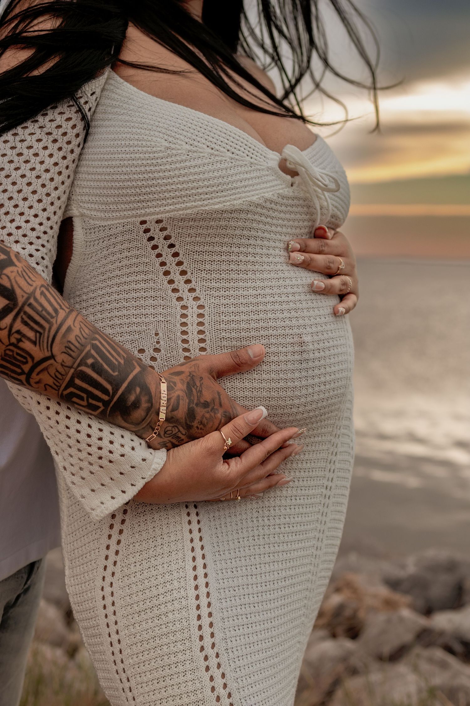 Pregnant woman in white knit dress cradling baby bump with tattooed partner hands at sunset beach.