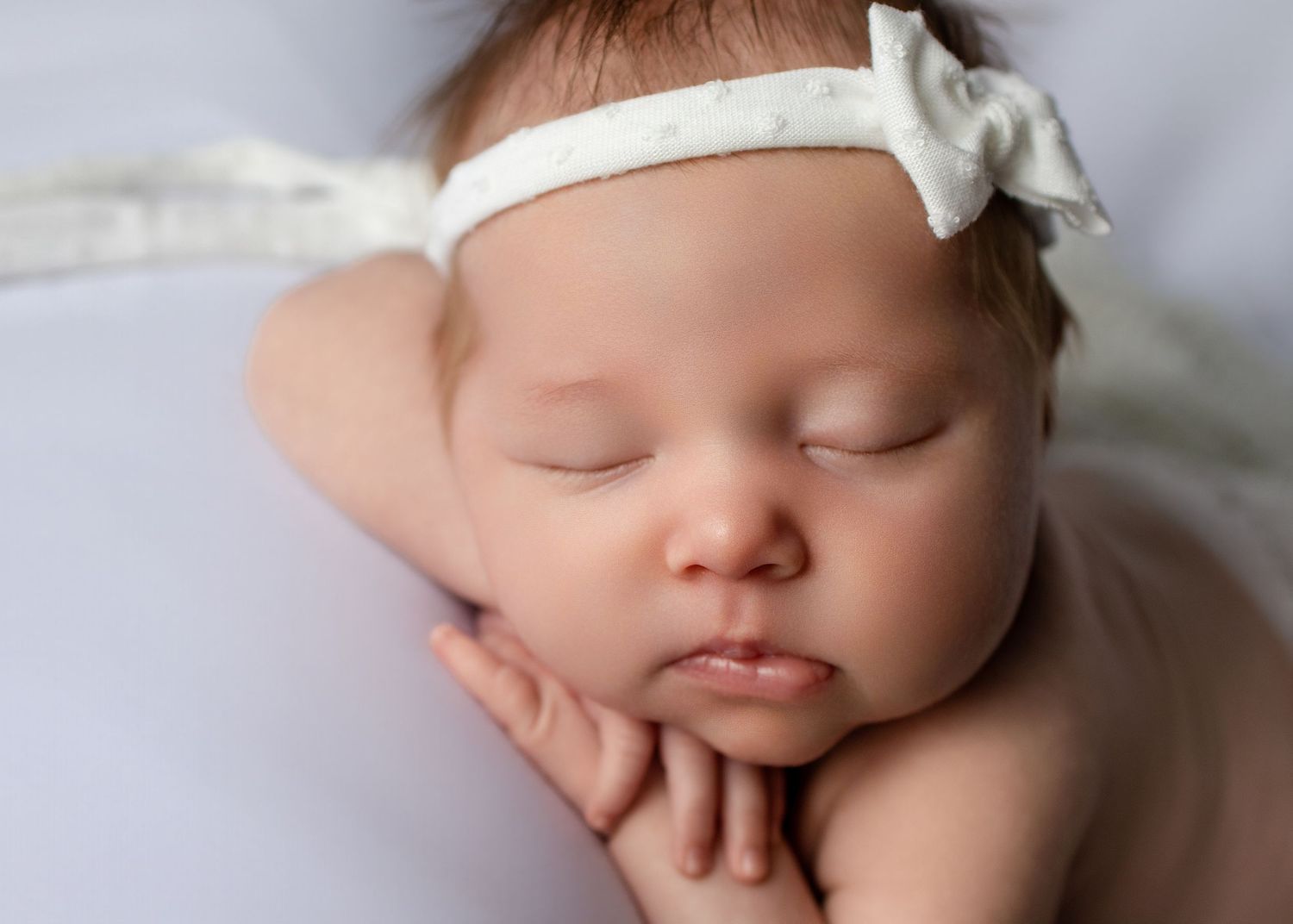 Sleeping newborn baby girl wearing a white bow headband, resting her chin on her hands in a peaceful pose.