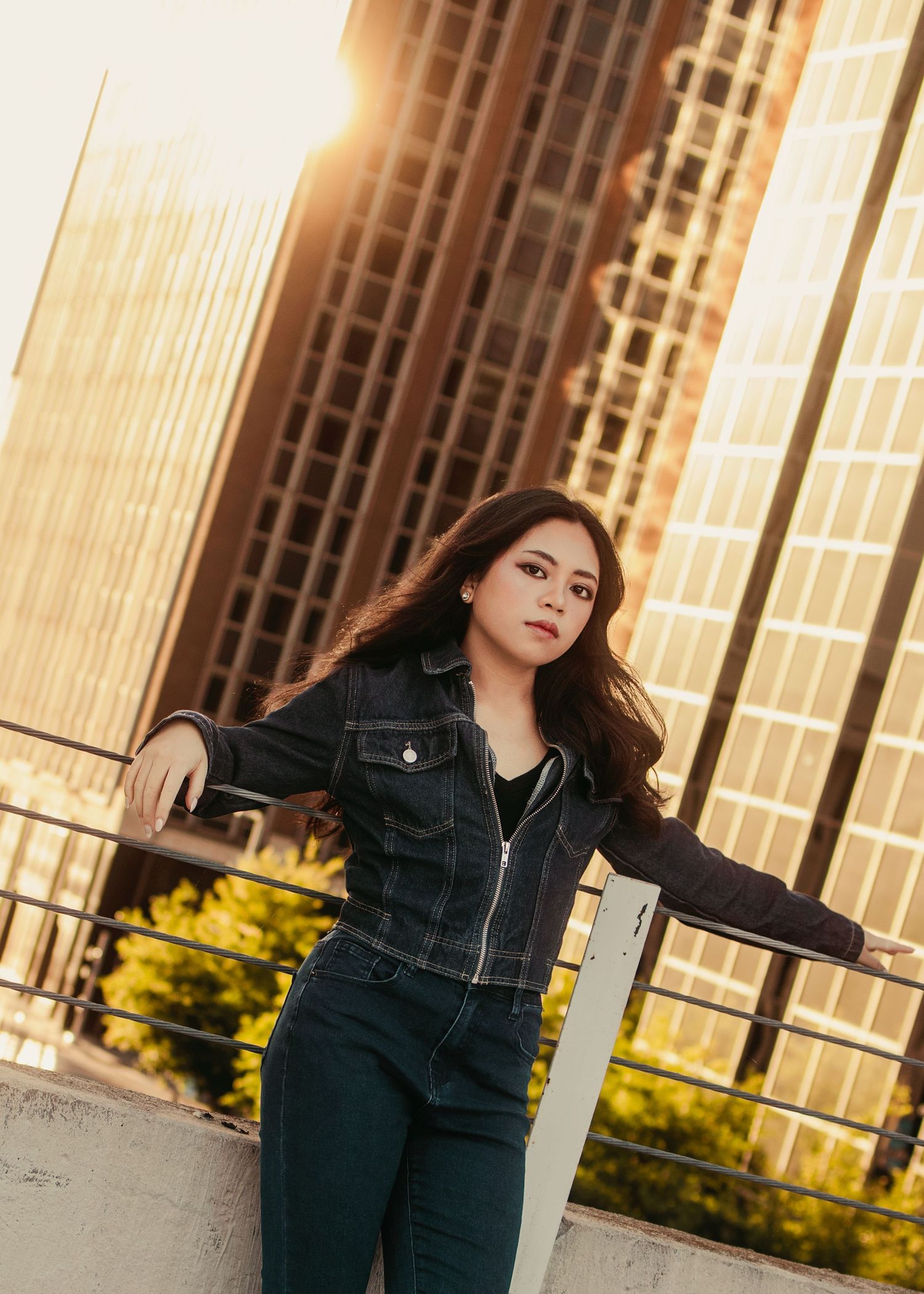 Young woman in dark denim jacket poses against city rooftop railing at golden hour sunset.