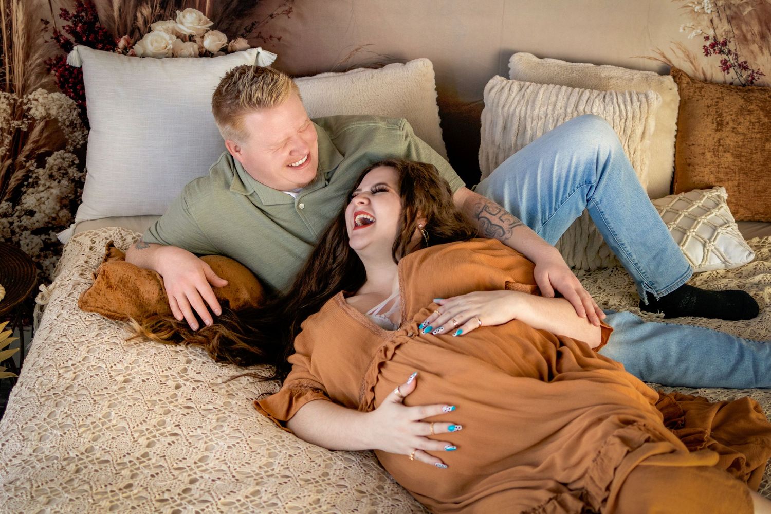 Laughing couple during maternity photo shoot, lying on a bed with warm earth tones, woman cradling baby bump.