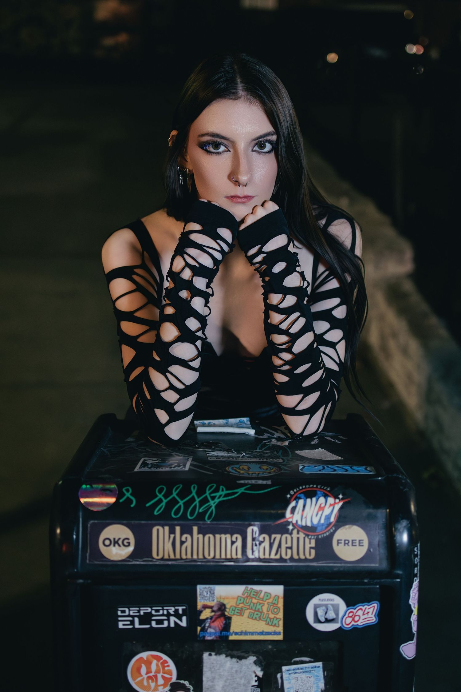 Young woman in black cutout dress leans on Oklahoma Gazette box covered in stickers.