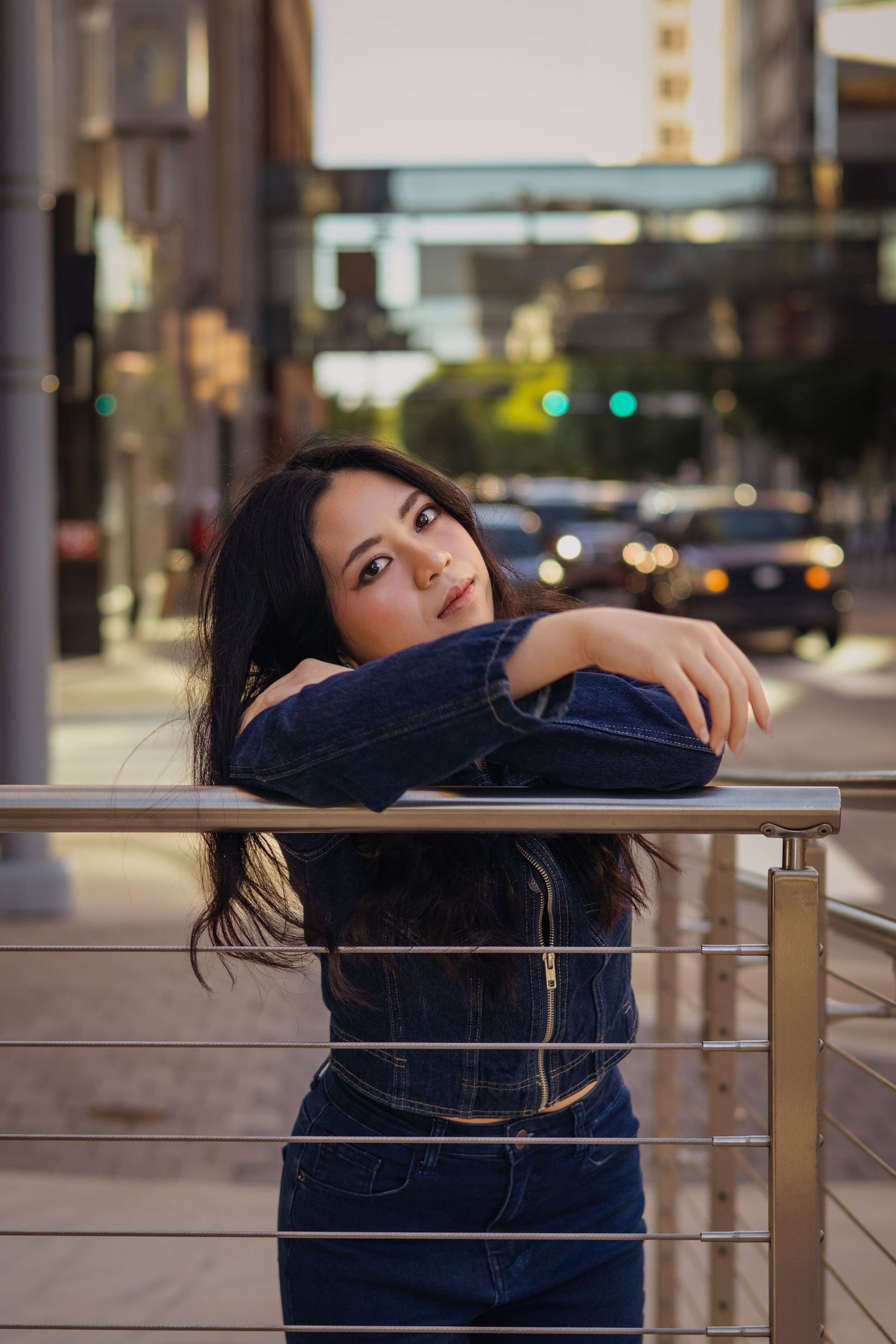 Young woman in denim jacket leaning on metal railing on city street at dusk with bokeh lights.