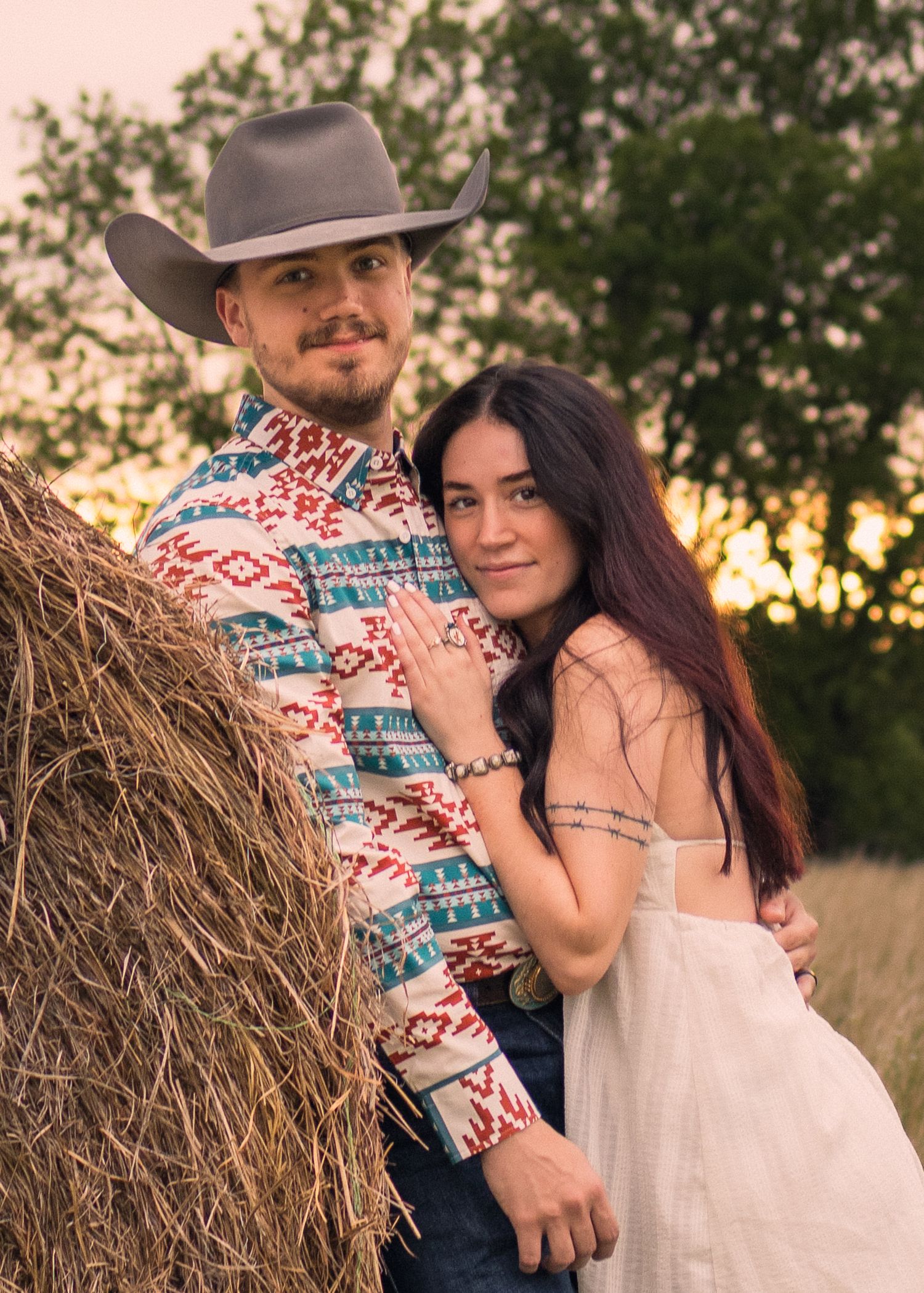 Couple posing by a hay bale at sunset, man in cowboy hat and southwestern shirt, woman in white dress.
