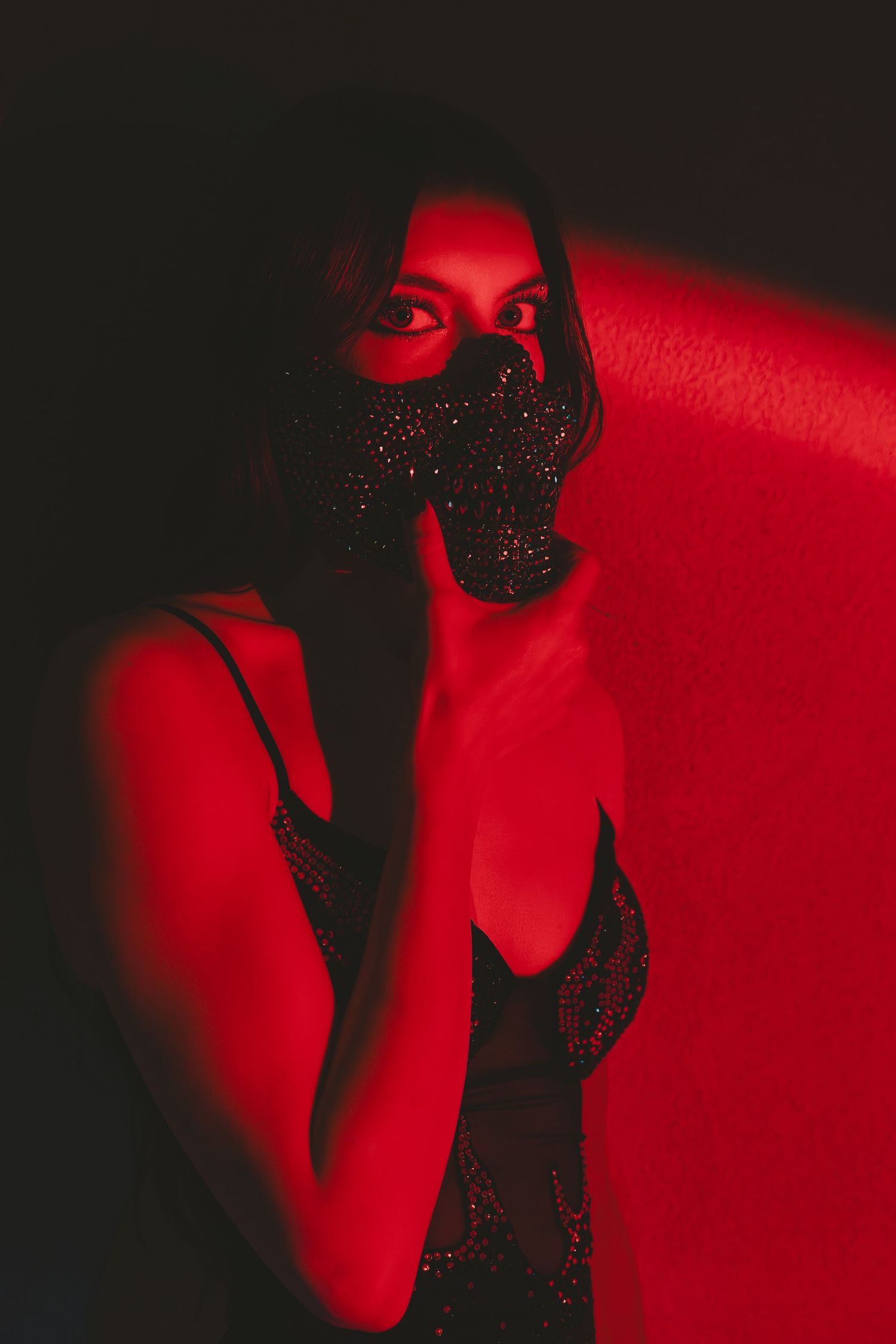 Woman in sparkly mask posed against red-lit wall wearing sequined dress in dramatic dark photography.