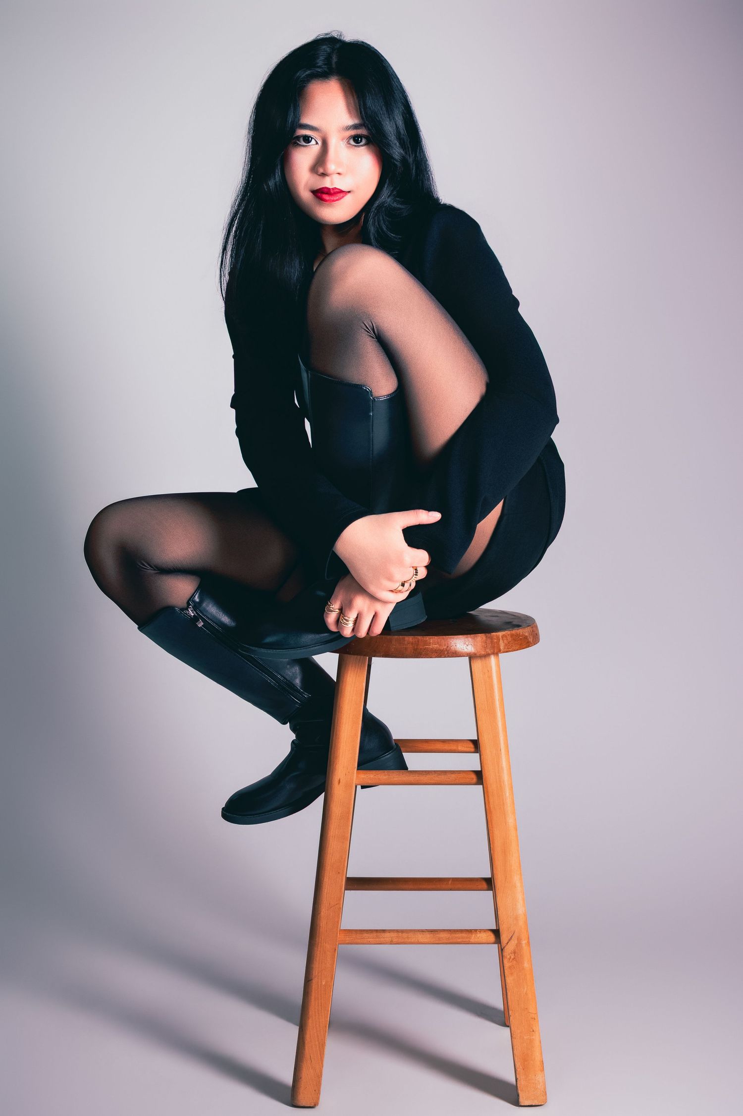 Young Asian woman in black dress and boots posing on wooden stool in photography studio.