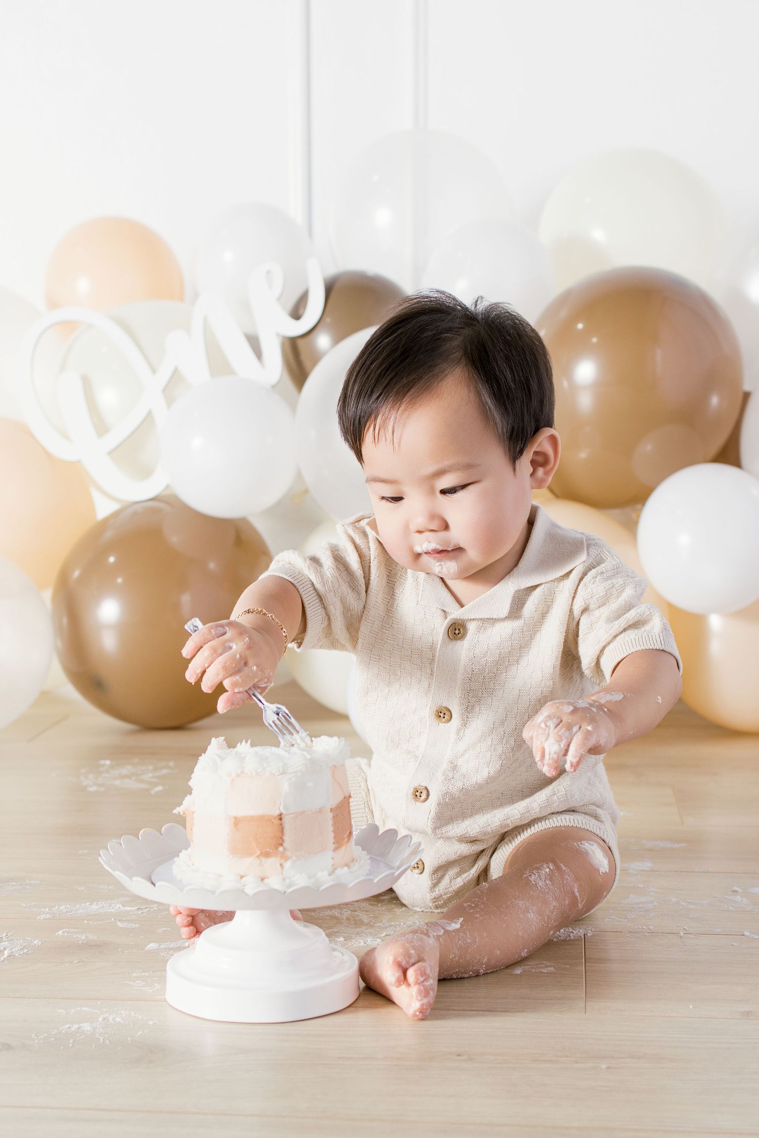 Baby boy in beige romper smashing first birthday cake surrounded by gold and white balloons with One sign.