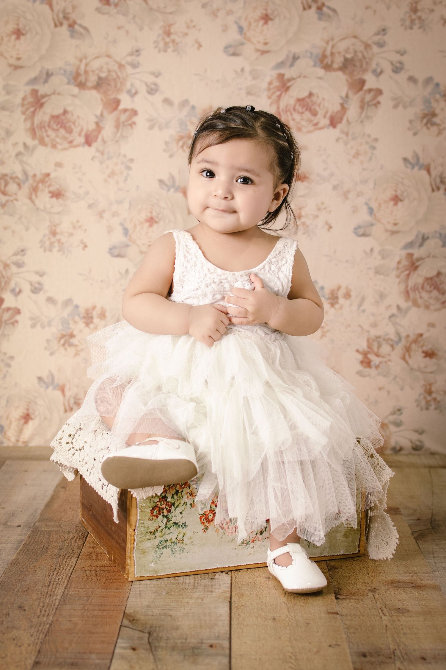 Baby girl in white tulle dress sitting on vintage floral box on wooden floor.