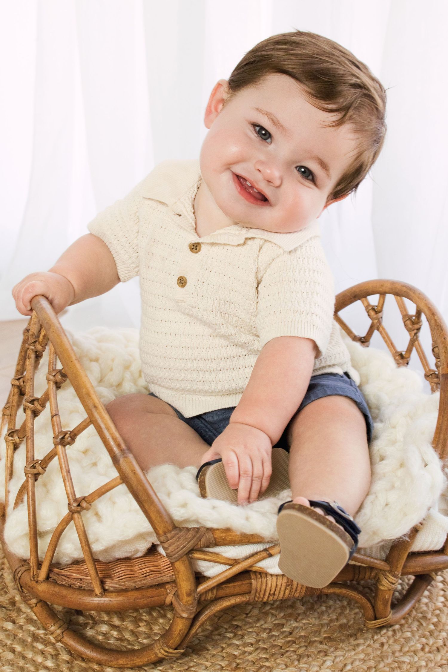 Smiling toddler boy in cream knit shirt and denim shorts sitting in a wicker rocking chair with a white blanket.